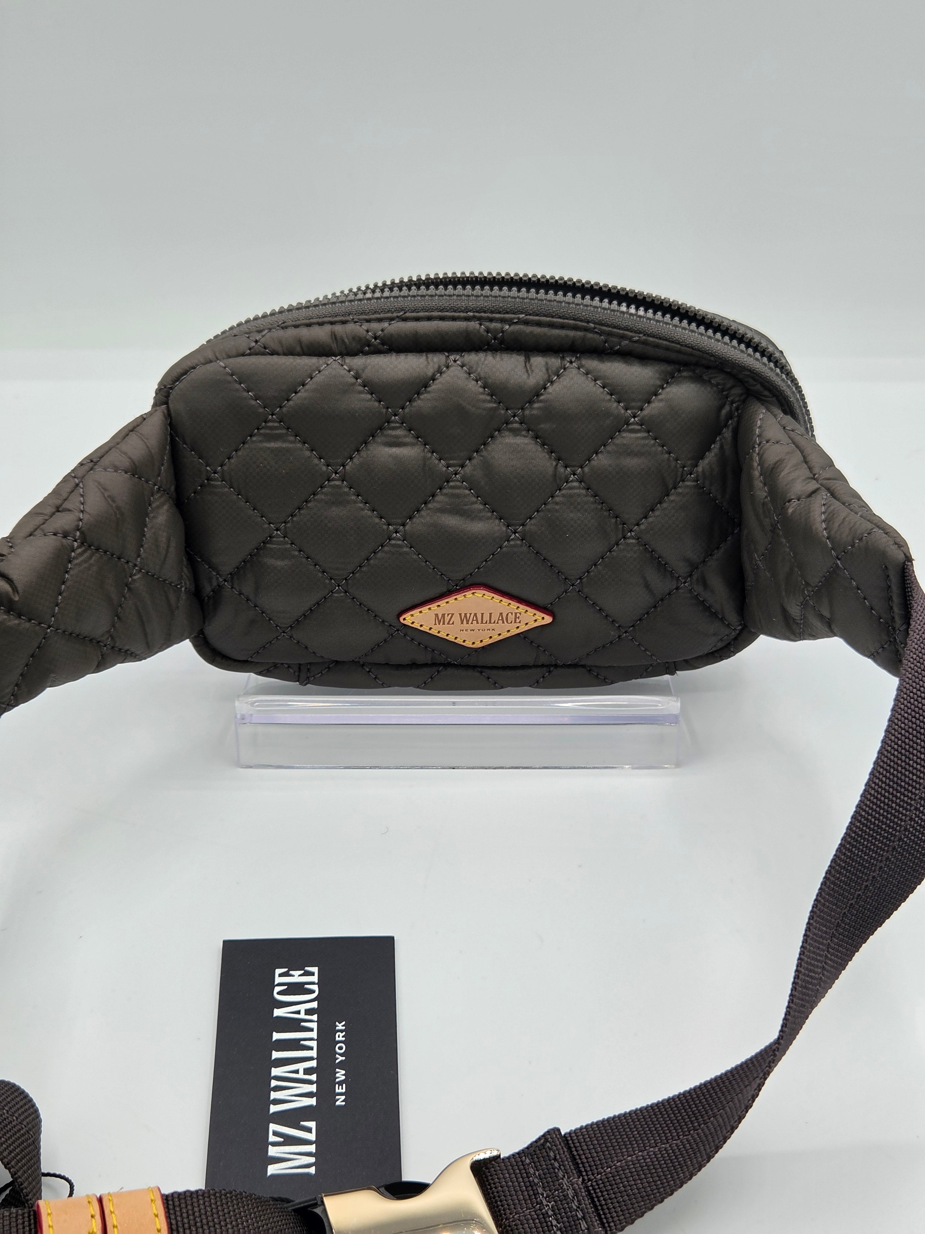 A dark brown quilted fanny pack with a diamond-stitched pattern, a small tan leather brand patch, and a black adjustable strap, displayed on a white surface with a brand card.