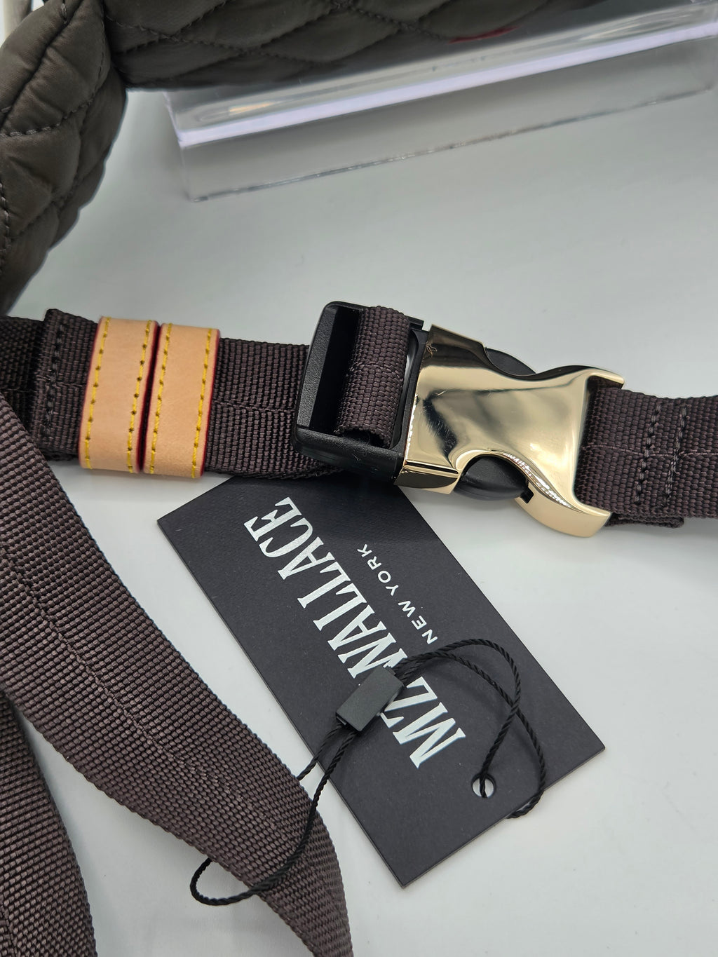 A close-up of a dark brown woven strap with a gold-tone metal buckle, a black plastic quick-release slider, and a tan leather accent tab. A black brand tag reading 'MZ MILAGE NEW YORK' is attached with a black string, resting on a light gray surface.