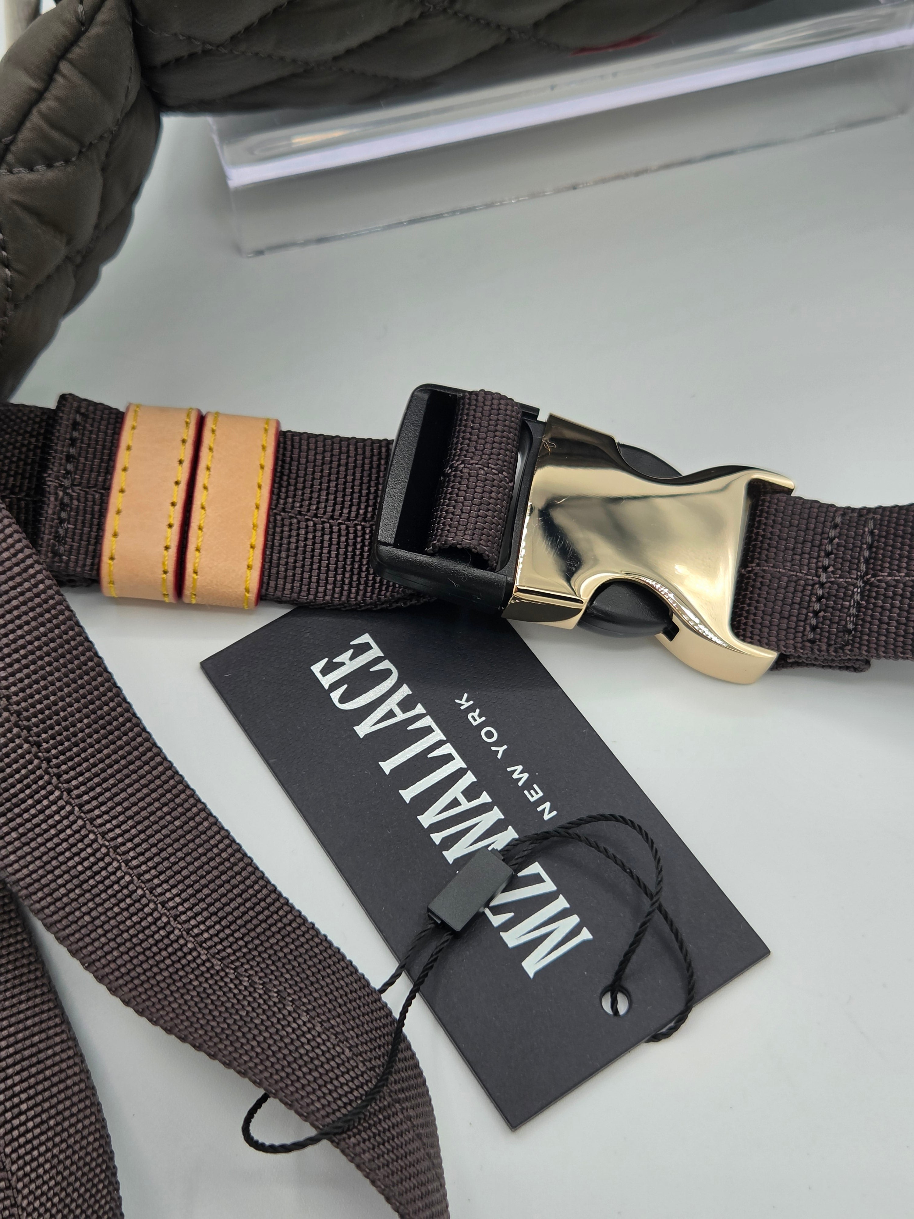 A close-up of a dark brown woven strap with a gold-tone metal buckle, a black plastic quick-release slider, and a tan leather accent tab. A black brand tag reading 'MZ MILAGE NEW YORK' is attached with a black string, resting on a light gray surface.