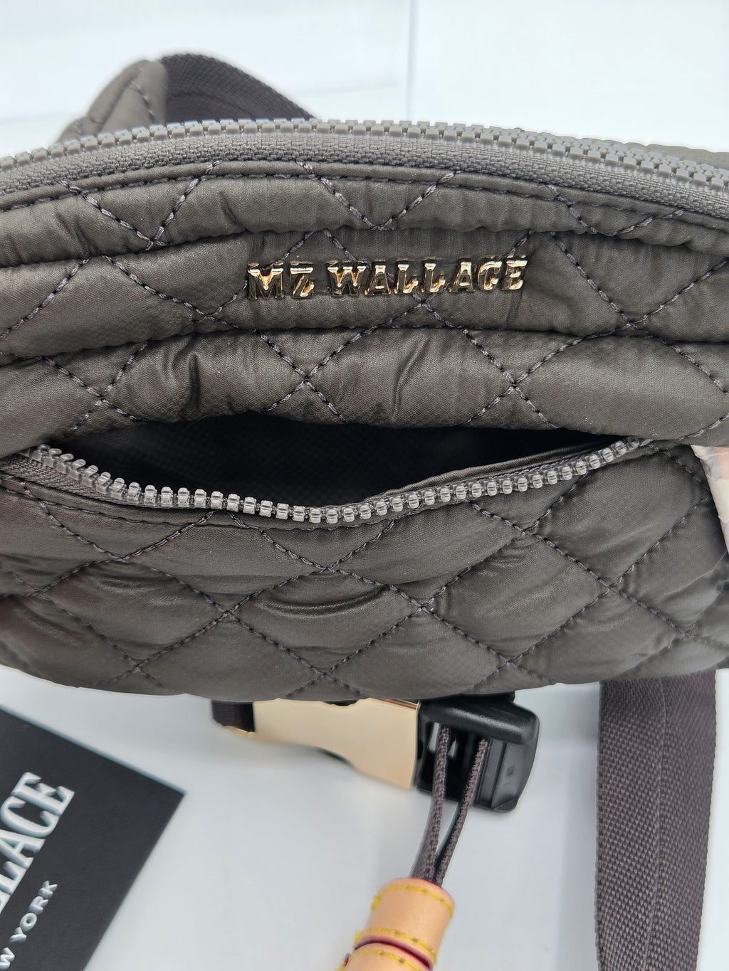 Close-up of a quilted gray shoulder bag with a gold-tone 'MZ WALLACE' logo on the front, showing the interior zip pocket and strap attachment.