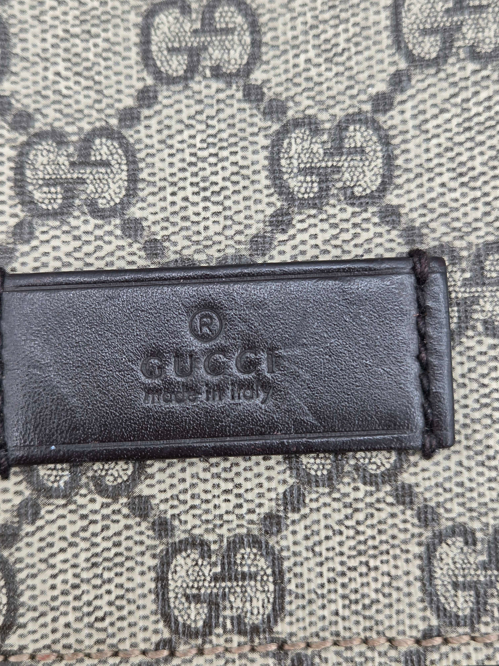 Close-up of a Gucci leather label embossed with 'GUCCI made in Italy' on a beige and gray GG Supreme canvas background.