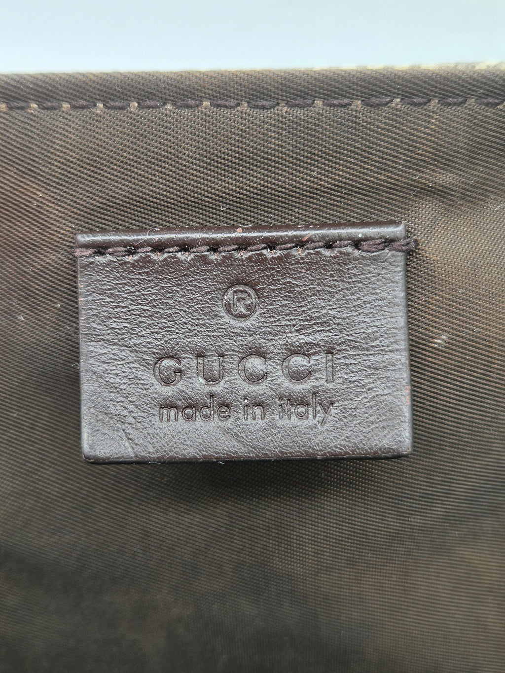 Close-up of a Gucci leather label embossed with 'GUCCI made in Italy' on a dark olive green woven fabric background.