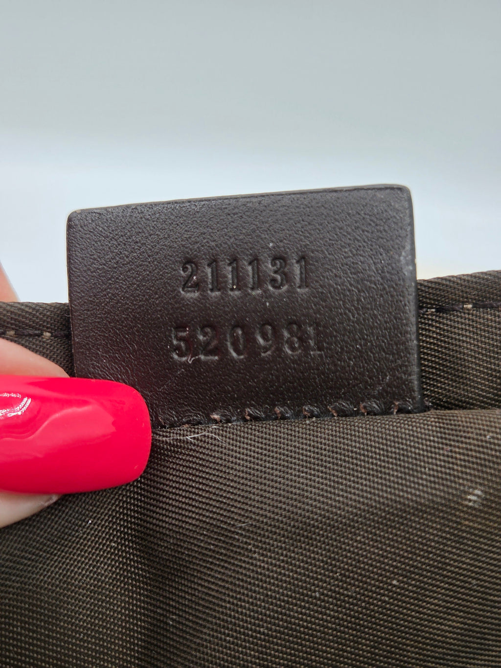 Close-up of a dark brown leather interior label stamped with embossed numbers '211131 520981' on a brown woven fabric surface, with a hand holding the edge.