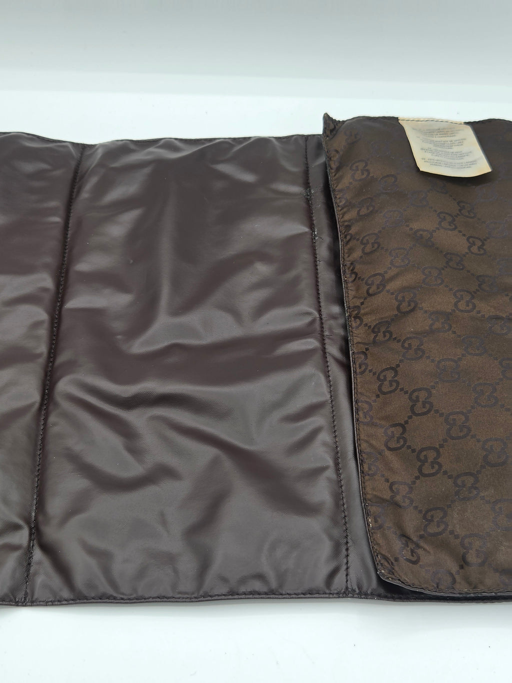 A brown Gucci foldable pouch or sleeve, shown open on a white background. The left side features a smooth dark brown satin-like interior, while the right side displays the classic brown GG Supreme monogram canvas exterior with a small beige paper tag attached.