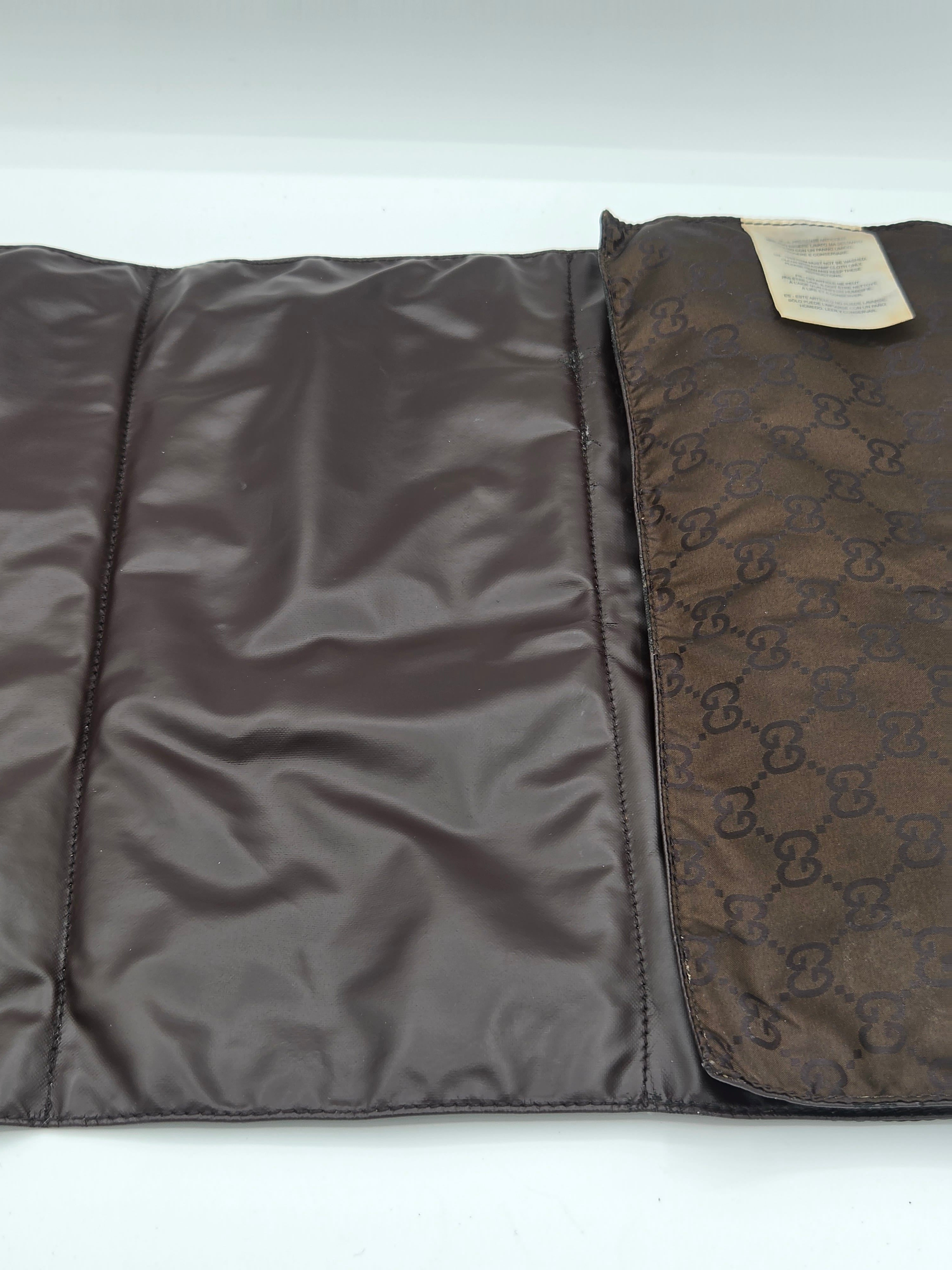 A brown Gucci foldable pouch or sleeve, shown open on a white background. The left side features a smooth dark brown satin-like interior, while the right side displays the classic brown GG Supreme monogram canvas exterior with a small beige paper tag attached.