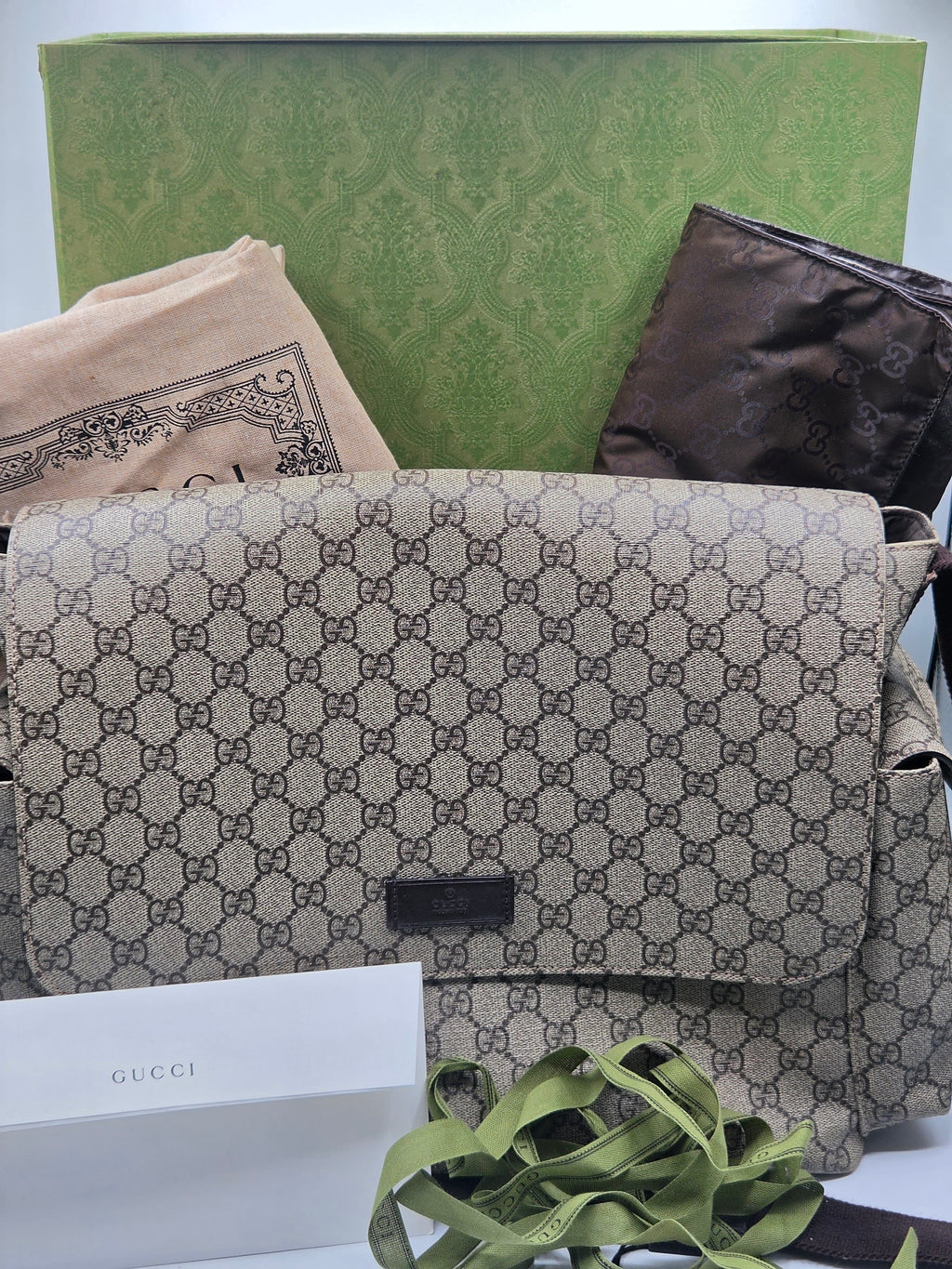 Gucci shoulder bag in beige and brown GG Supreme canvas, shown with its green dust bag, brown pouch, white Gucci box, and green ribbon on a white surface.