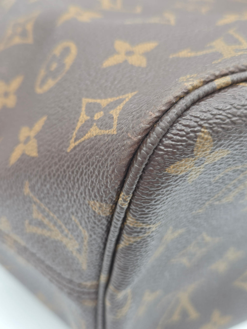 Close-up of a Louis Vuitton monogram canvas bag corner showing the classic brown and tan LV pattern with pebbled leather texture and a curved seam.
