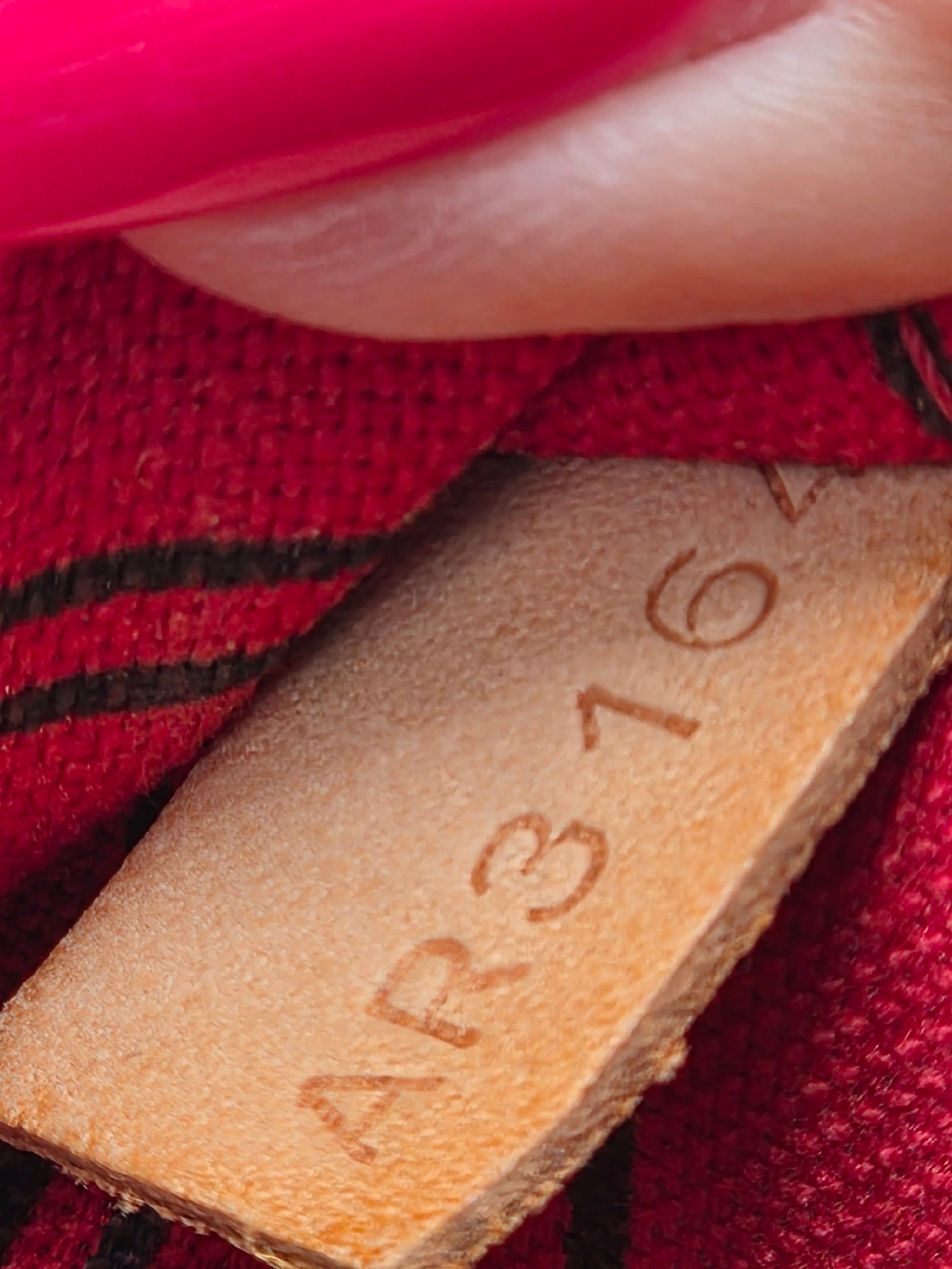 Close-up of a tan leather interior label stamped with the numbers AR3162, set against a red and black patterned fabric background with a pink object at the top edge.