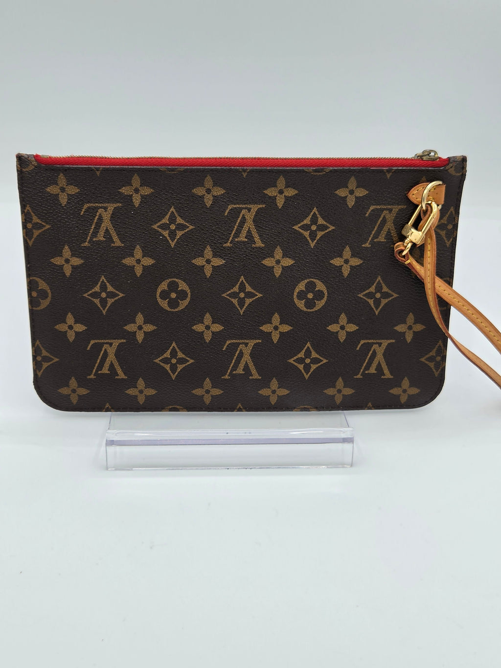 A Louis Vuitton monogram canvas wristlet pouch with a red zipper trim and a tan leather wristlet strap, displayed on a clear acrylic stand against a light gray background.