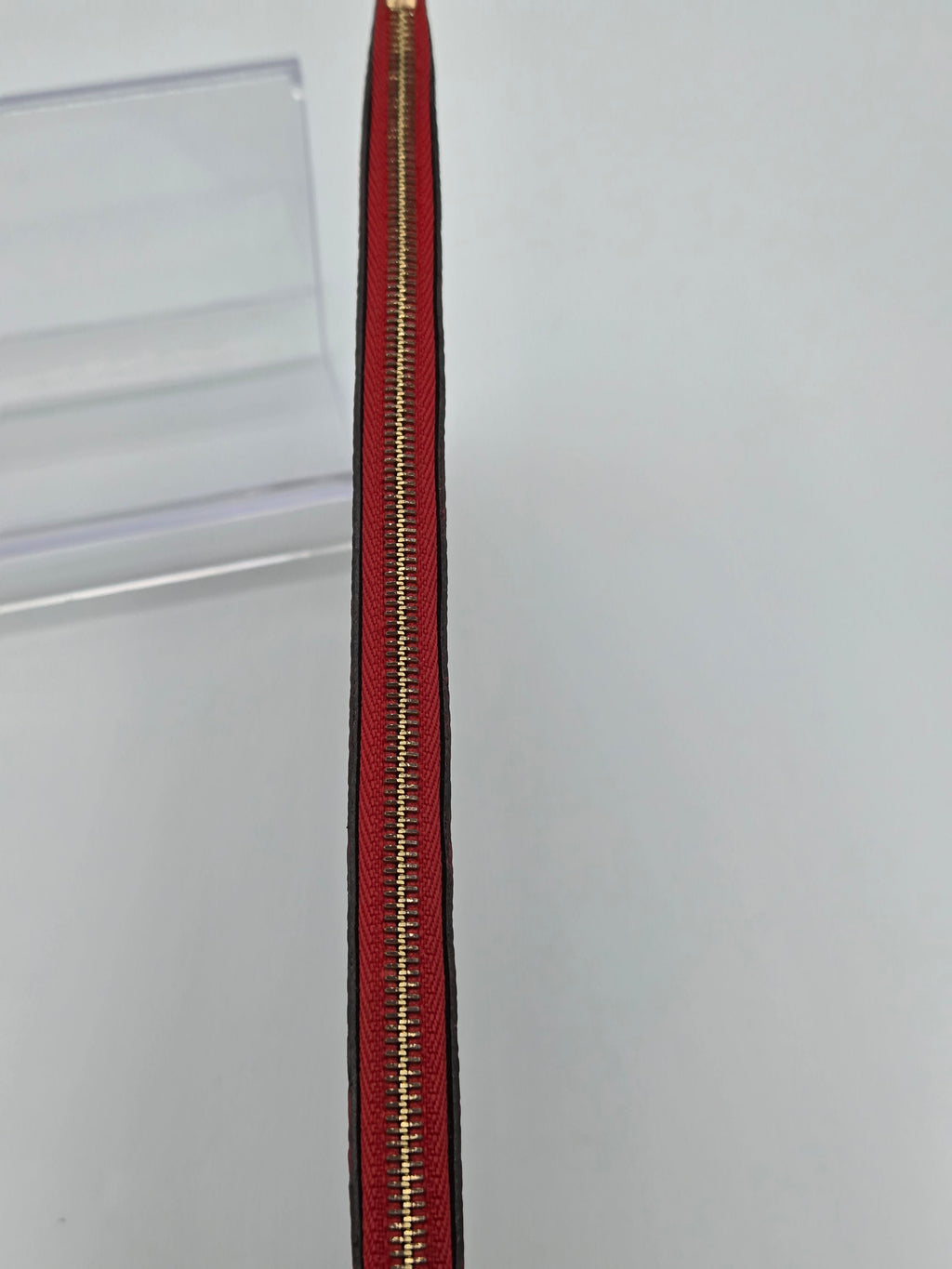 A close-up of a red zipper with gold metal teeth, shown vertically against a plain light gray background.