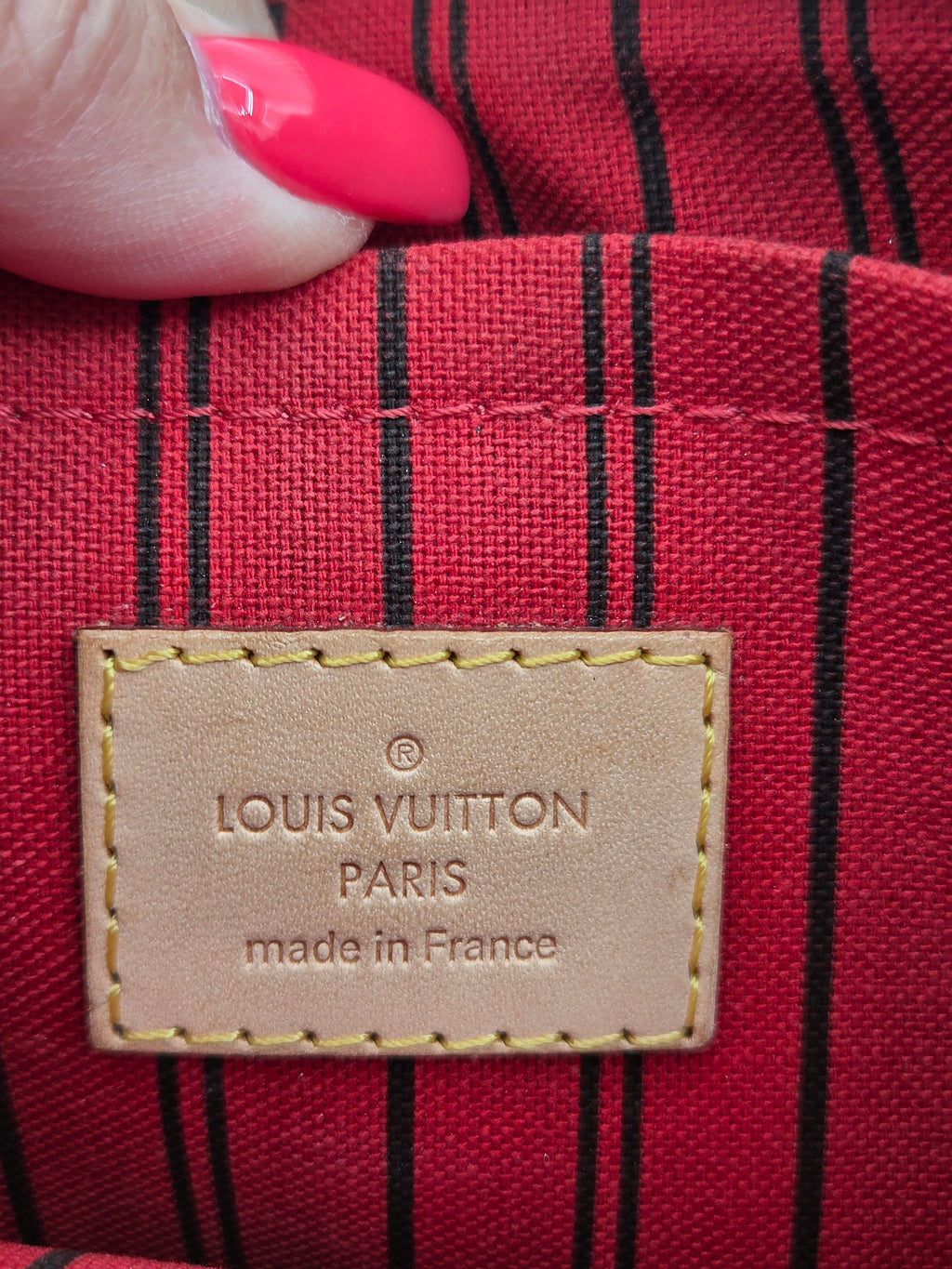 Close-up of a Louis Vuitton fabric interior with a tan leather brand patch reading 'LOUIS VUITTON PARIS made in France,' showing a red background with thin black vertical stripes and a finger with bright pink nail polish at the top edge.