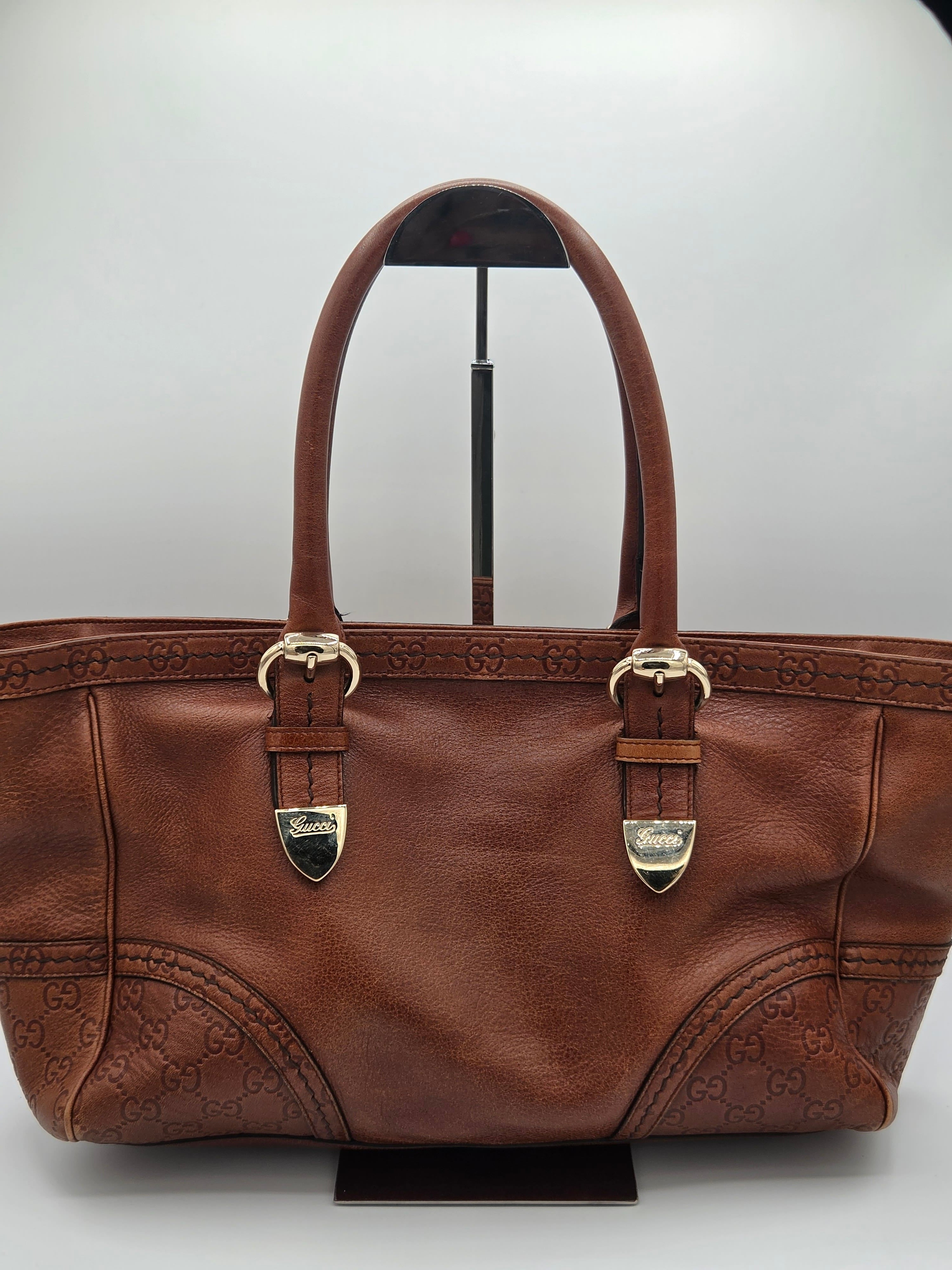 A brown leather handbag with embossed monogram pattern, dual top handles, and gold-tone hardware, displayed on a stand against a light gray background.