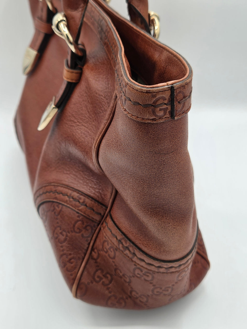 A close-up of a brown leather handbag showing the side and bottom corner, featuring embossed Gucci monogram pattern and gold-tone hardware.