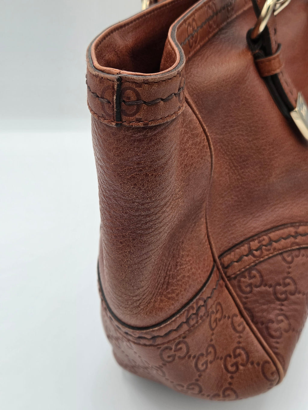 Close-up of a brown leather handbag showing the embossed Gucci GG monogram pattern, stitching, and part of a gold-tone metal clasp on a light gray background.