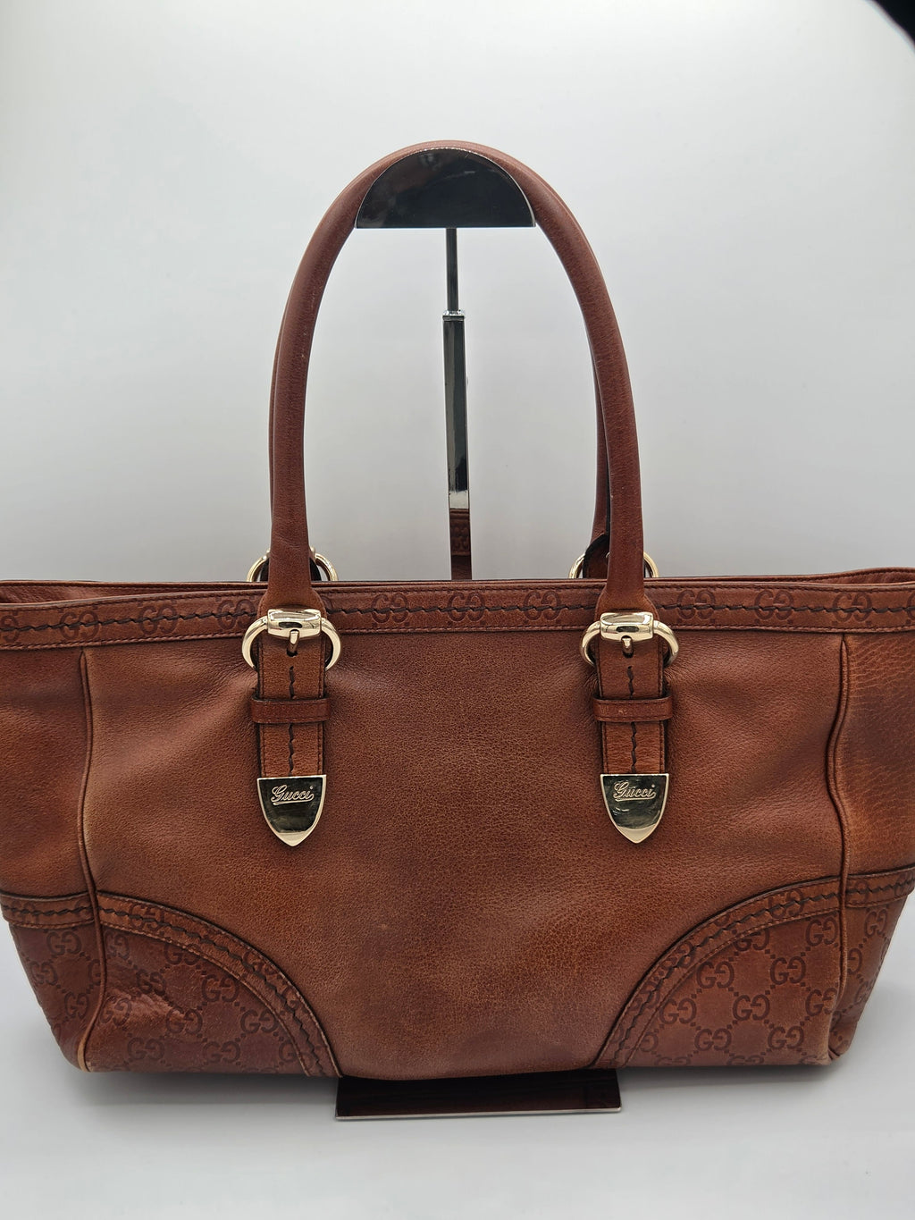 A brown leather handbag with embossed monogram pattern on the lower front panels and double top handles, displayed on a stand against a light gray background.