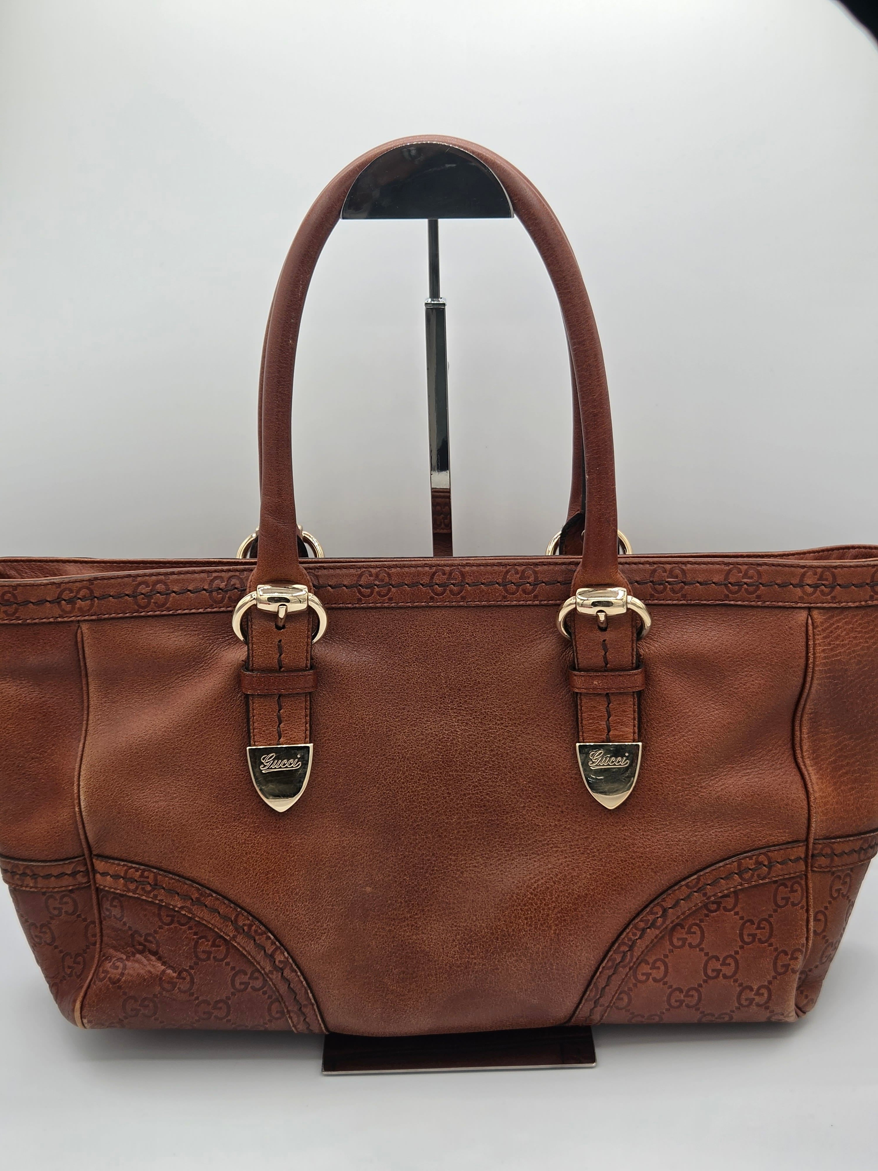 A brown leather handbag with embossed monogram pattern on the lower front panels and double top handles, displayed on a stand against a light gray background.