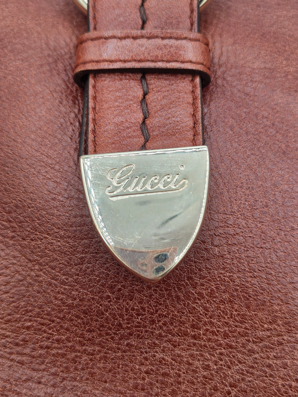 Close-up of a brown leather strap with a silver-tone shield-shaped Gucci buckle on a textured brown leather surface.