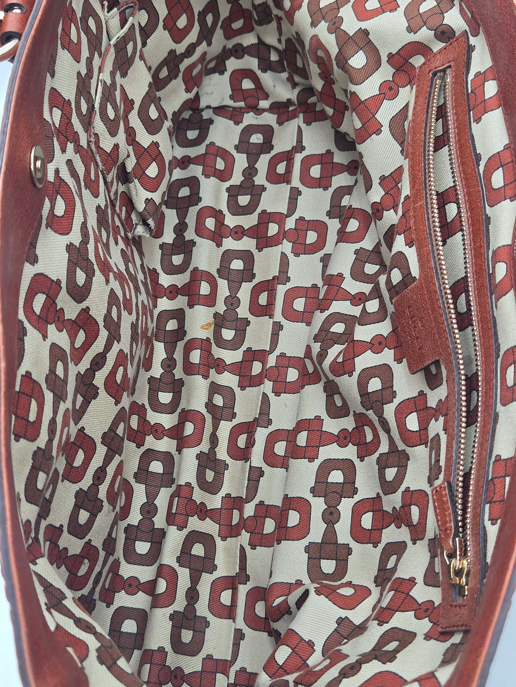 Close-up of the interior lining of a leather bag, showing a repeating monogram-style pattern in red and dark brown on a light beige background, with a zippered pocket on the right side.