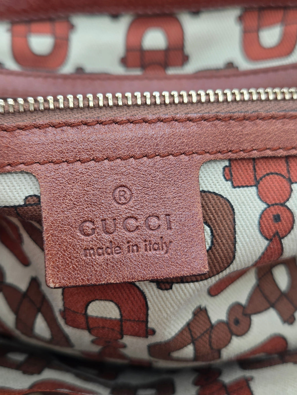 Close-up of a Gucci zippered pouch featuring a brown leather patch with 'GUCCI made in italy' embossed, set against a beige canvas background with a brown and red repeating pattern.
