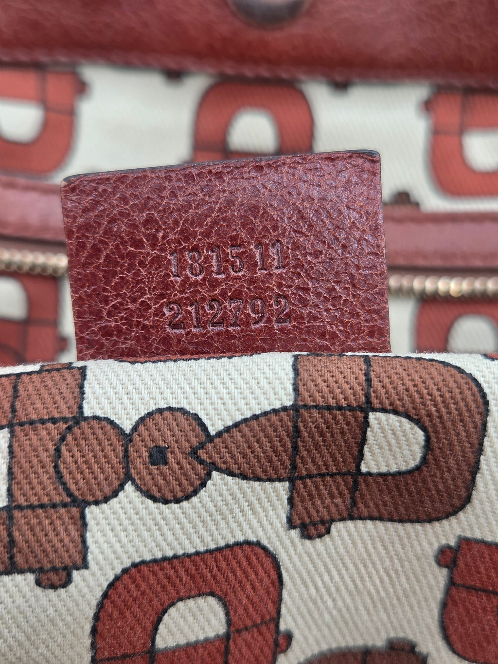 Close-up of a canvas pouch with a brown and red graphic pattern, featuring a brown leather tab stamped with embossed numbers 181511 and 212792.