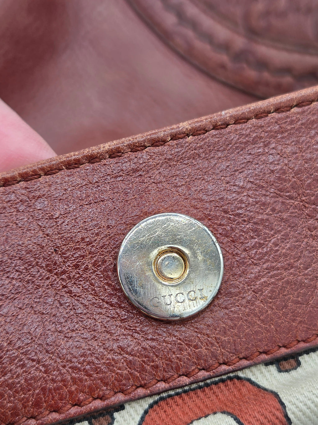 Close-up of a brown leather snap button closure with a visible Gucci logo stamped on the metal, alongside a hint of the brand's patterned fabric.