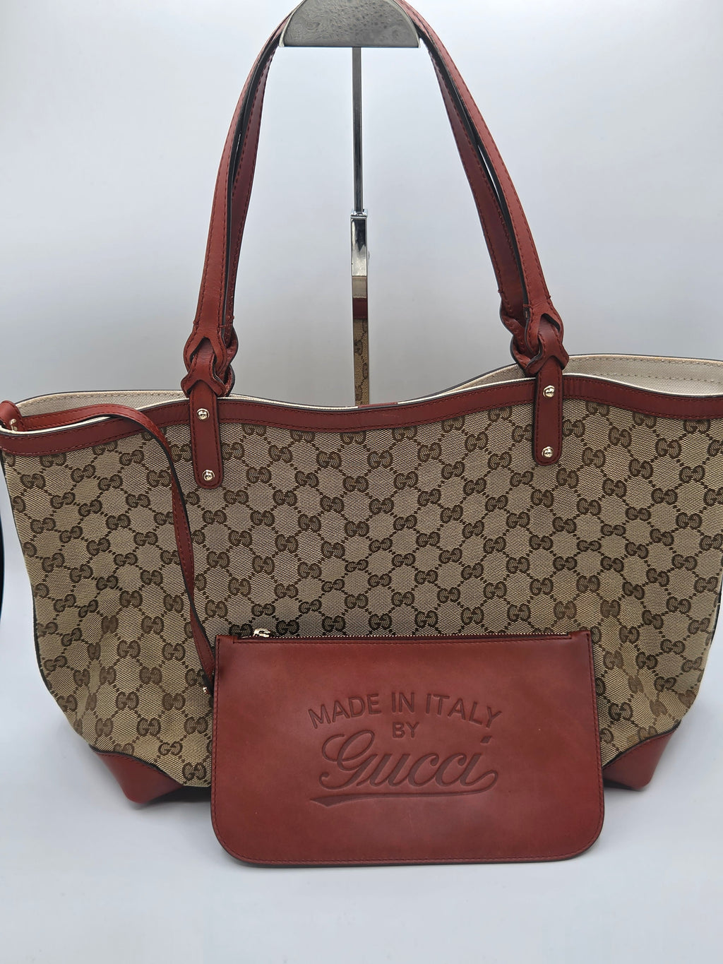 Gucci monogram canvas tote bag with brown leather handles and trim, shown with a matching brown leather pouch in front on a white studio background.