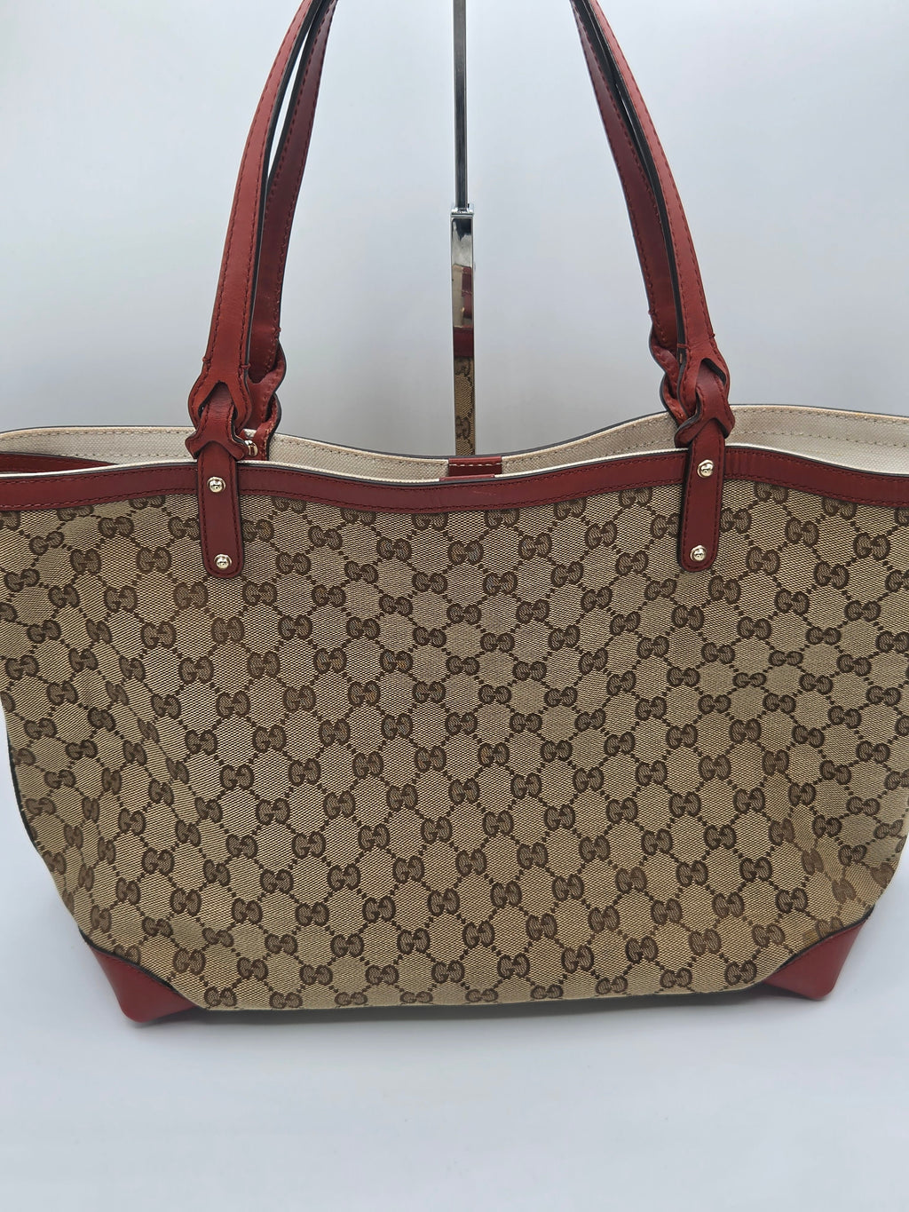 A vintage-style Gucci tote bag featuring the classic brown and beige GG monogram canvas, brown leather handles, and brown leather trim, displayed on a stand against a plain light gray background.