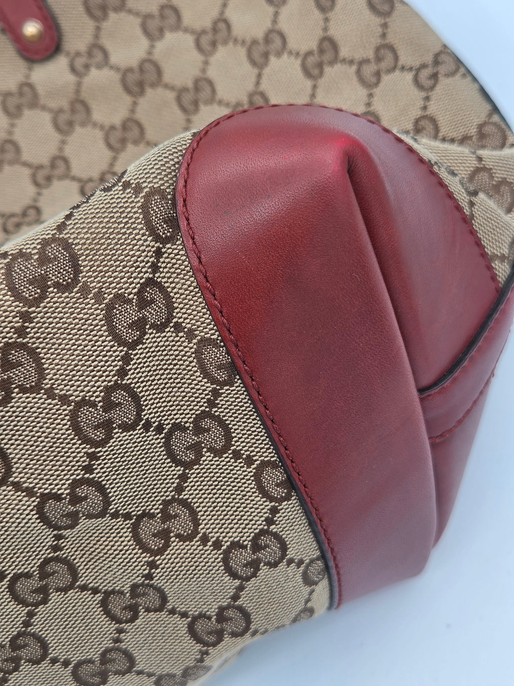 Close-up of a Gucci handbag corner showing beige GG monogram canvas with dark brown print and a prominent reddish-brown leather trim on a white background.