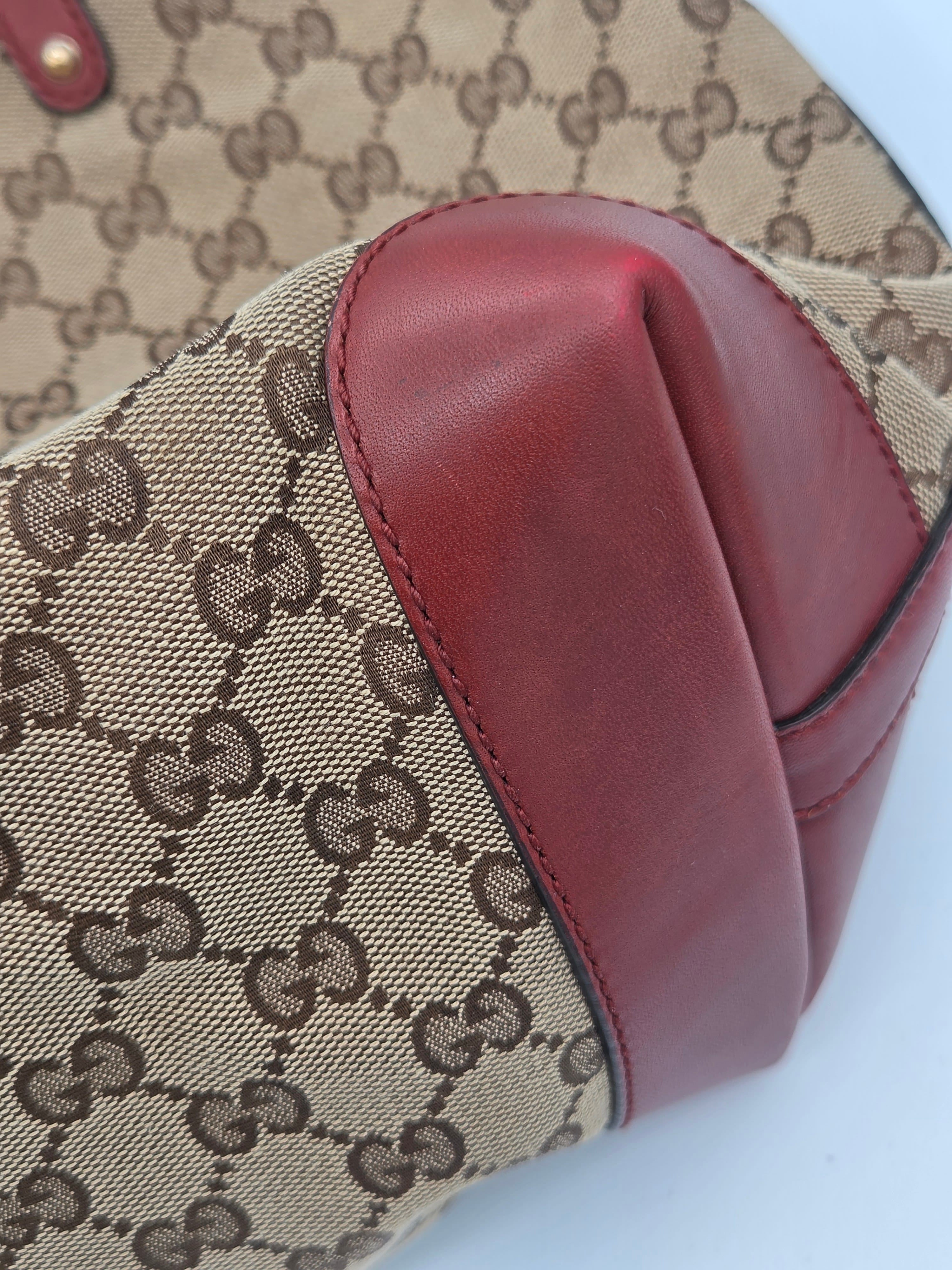 Close-up of a Gucci handbag corner showing beige GG monogram canvas with dark brown print and a prominent reddish-brown leather trim on a white background.