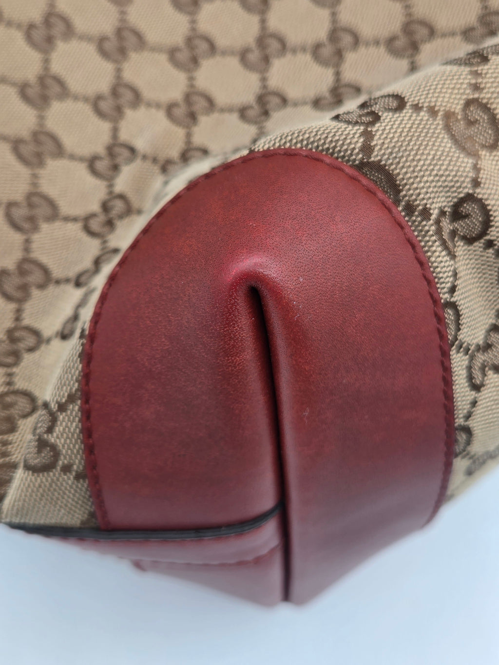 Close-up of a Gucci monogram canvas bag corner with a curved dark red leather trim and a black Gucci logo pattern on a beige background.