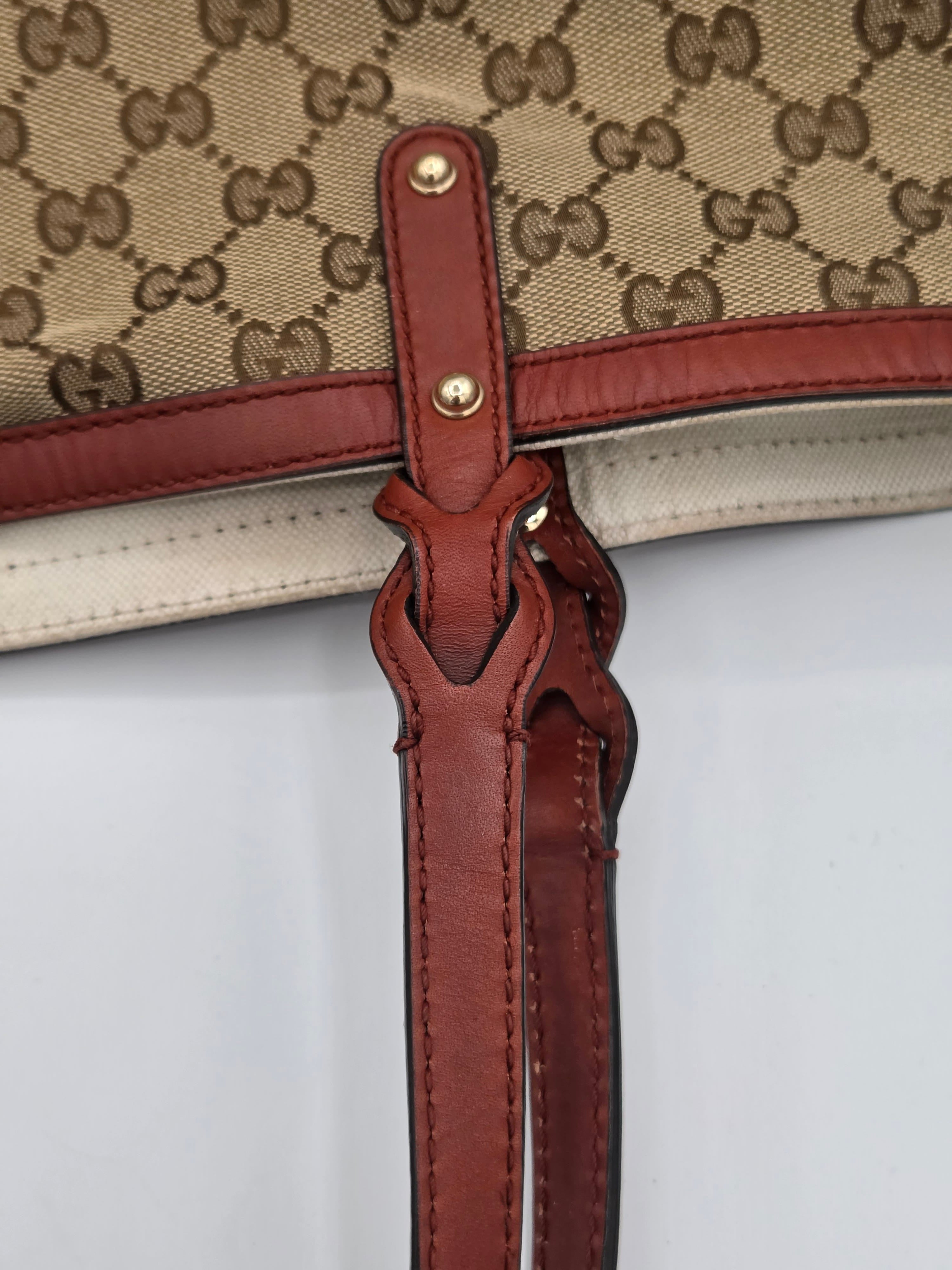 Close-up detail of a Gucci handbag showing the GG Supreme canvas, reddish-brown leather trim, and a leather strap with gold-tone hardware against a plain white background.