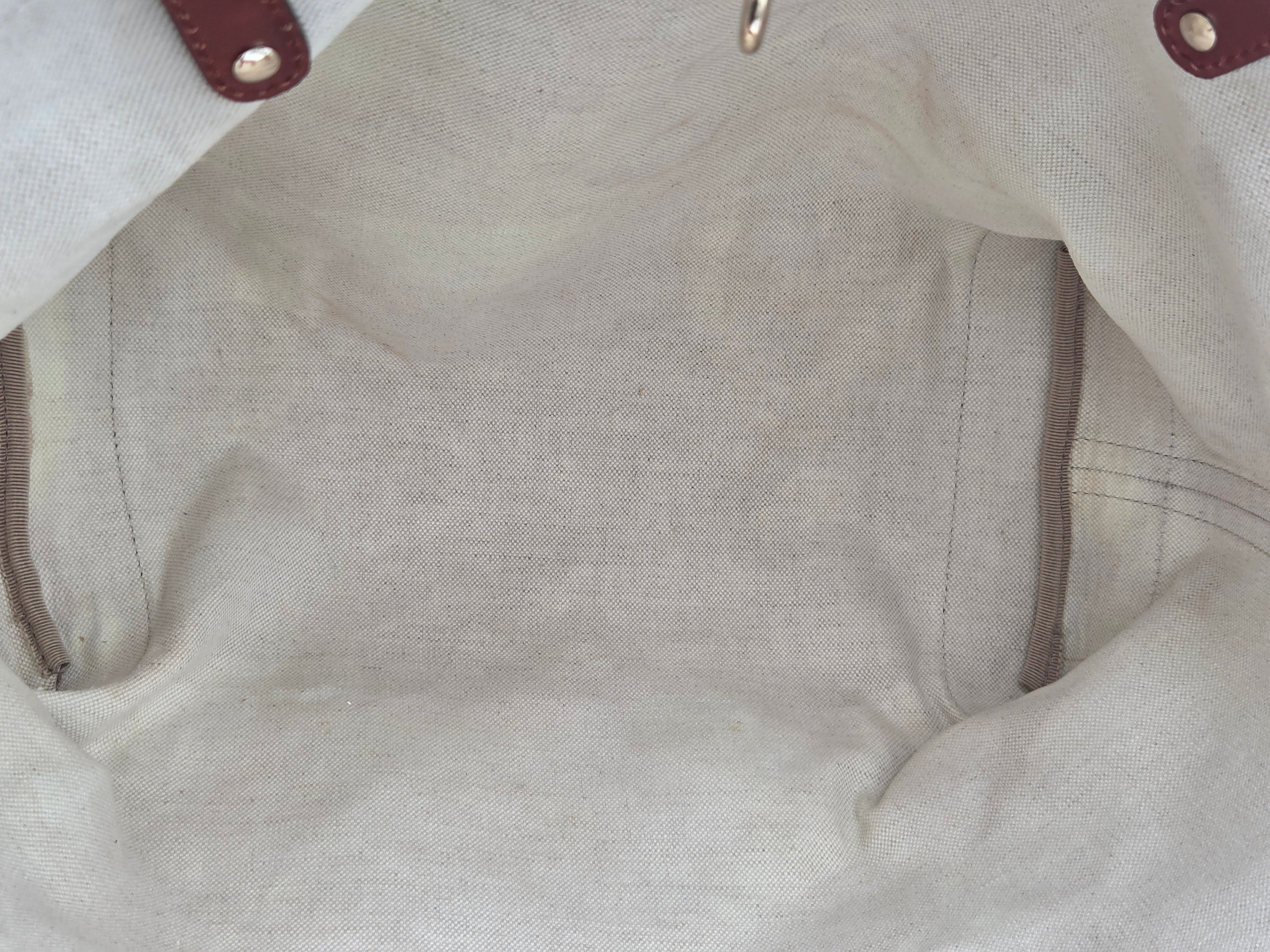 Close-up of a light beige canvas or linen fabric interior with visible stitching, a small metal ring near the top, and partial dark brown leather-like corner tabs with metal rivets.