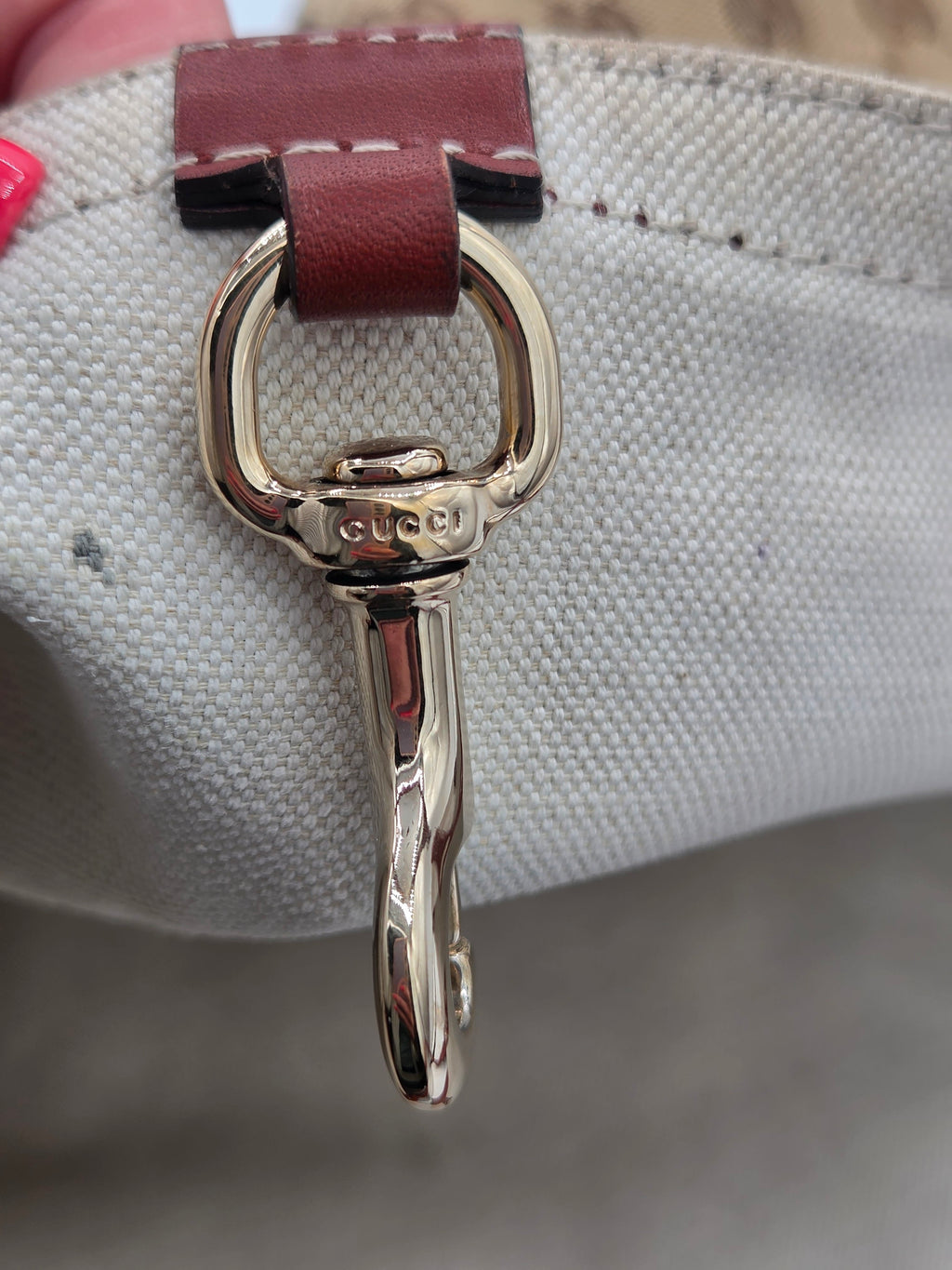 Close-up of a Gucci bag hardware detail featuring a polished gold-tone horsebit ring clasp engraved with the brand name, attached to a dark brown leather loop on a light beige canvas bag.
