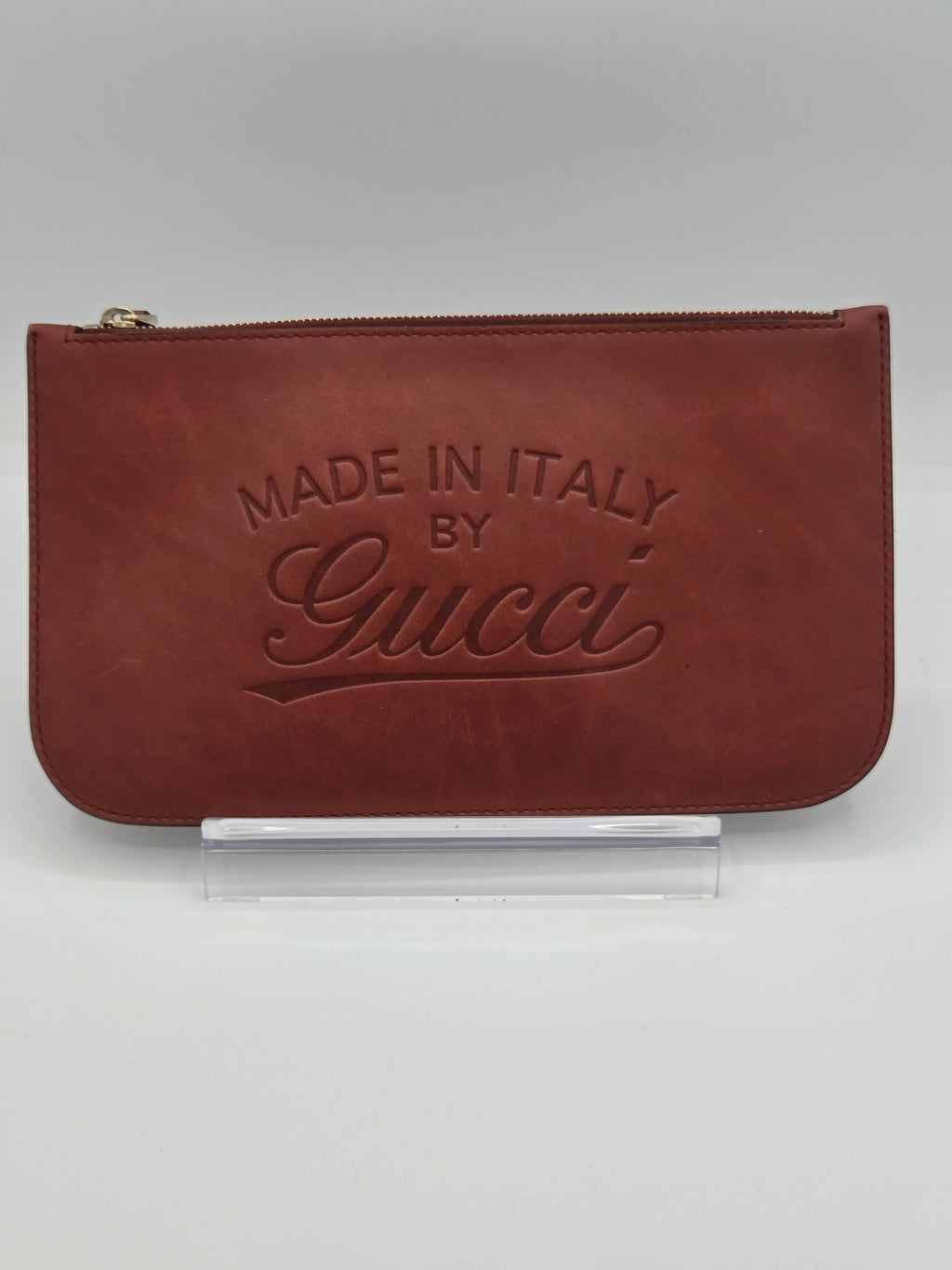 A reddish-brown leather zip pouch with an embossed 'Made in Italy by Gucci' logo, displayed on a clear acrylic stand against a neutral gray background.