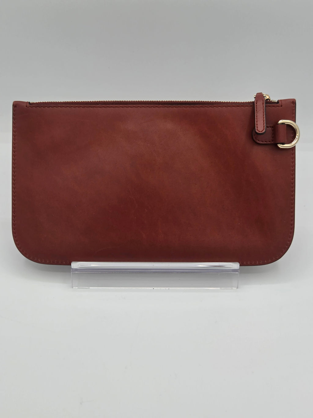 A reddish-brown leather zip-around pouch with a small leather tab and gold-tone ring detail on the right side, displayed upright on a clear acrylic stand against a light gray background.