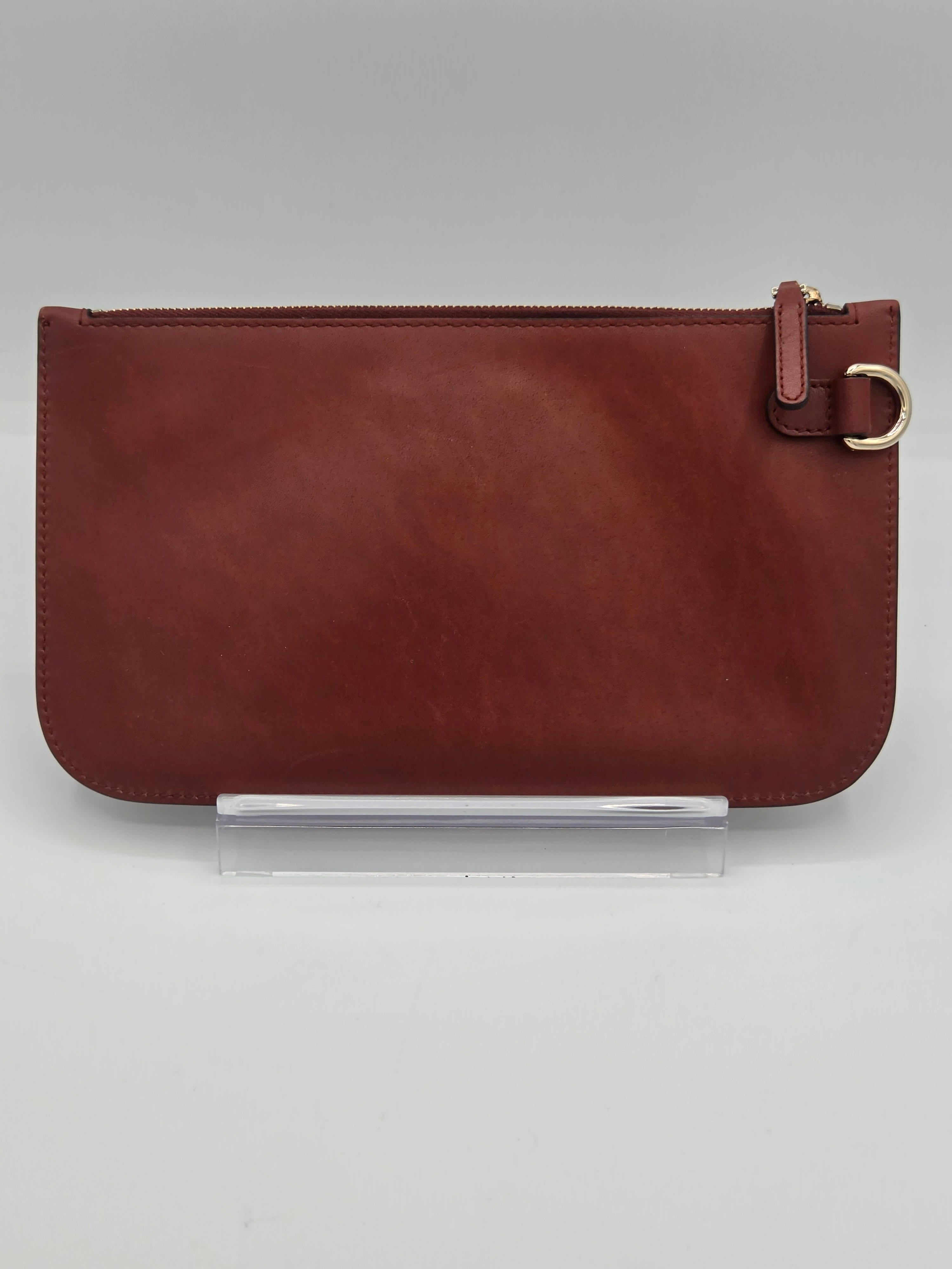 A reddish-brown leather zip-around pouch with a small leather tab and gold-tone ring detail on the right side, displayed upright on a clear acrylic stand against a light gray background.