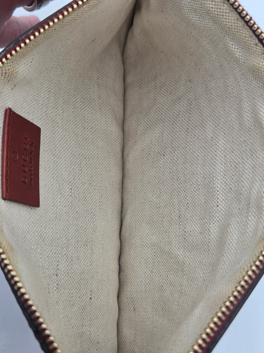 Close-up of the interior of a canvas zippered pouch or clutch, showing a beige woven fabric lining, a brown leather brand patch embossed with 'CLOTHING', and a gold-toned zipper.