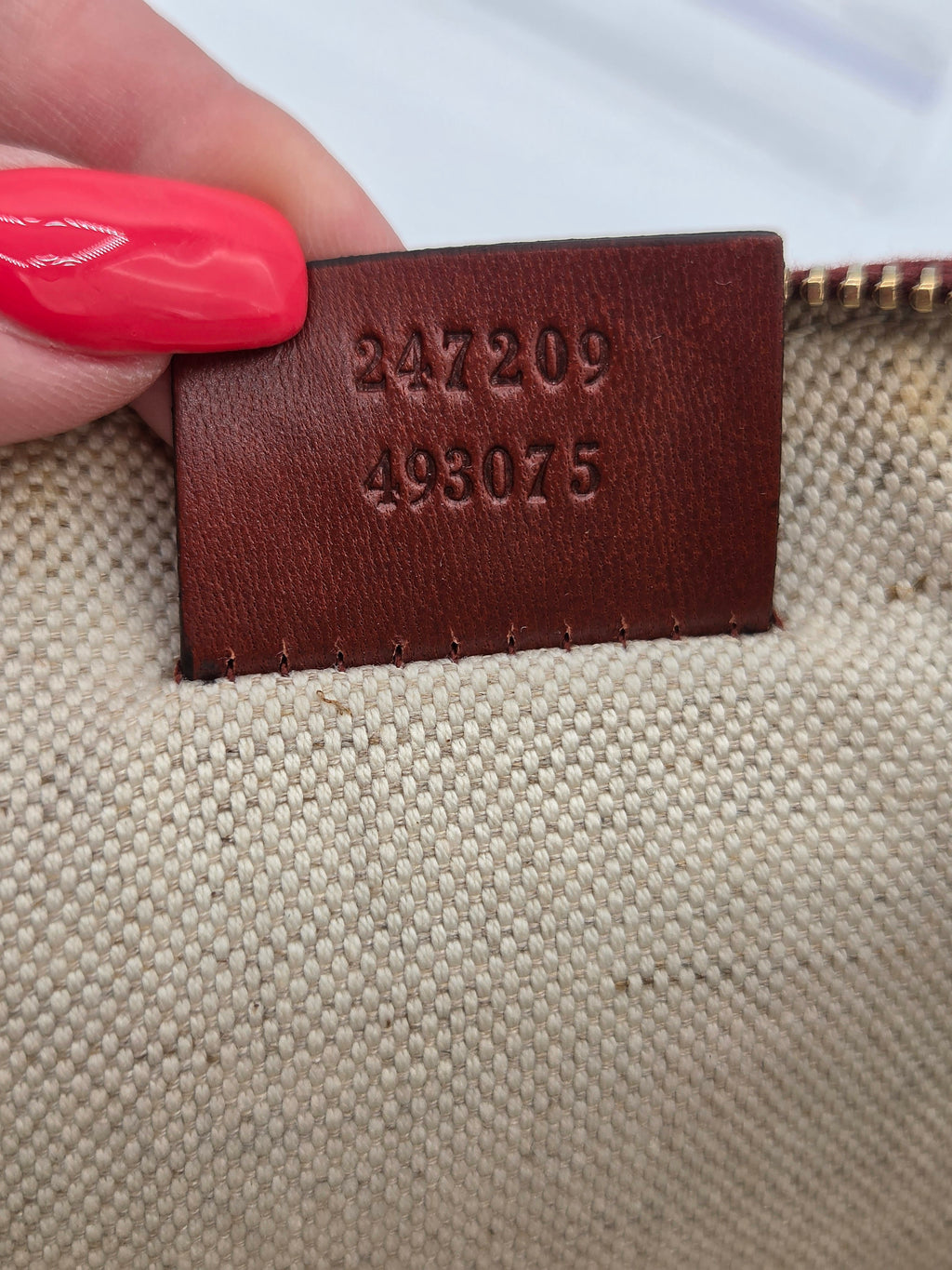 Close-up of a beige woven fabric item with a brown leather tag stamped with the numbers 247209 and 498075, held by a hand with bright pink nail polish.