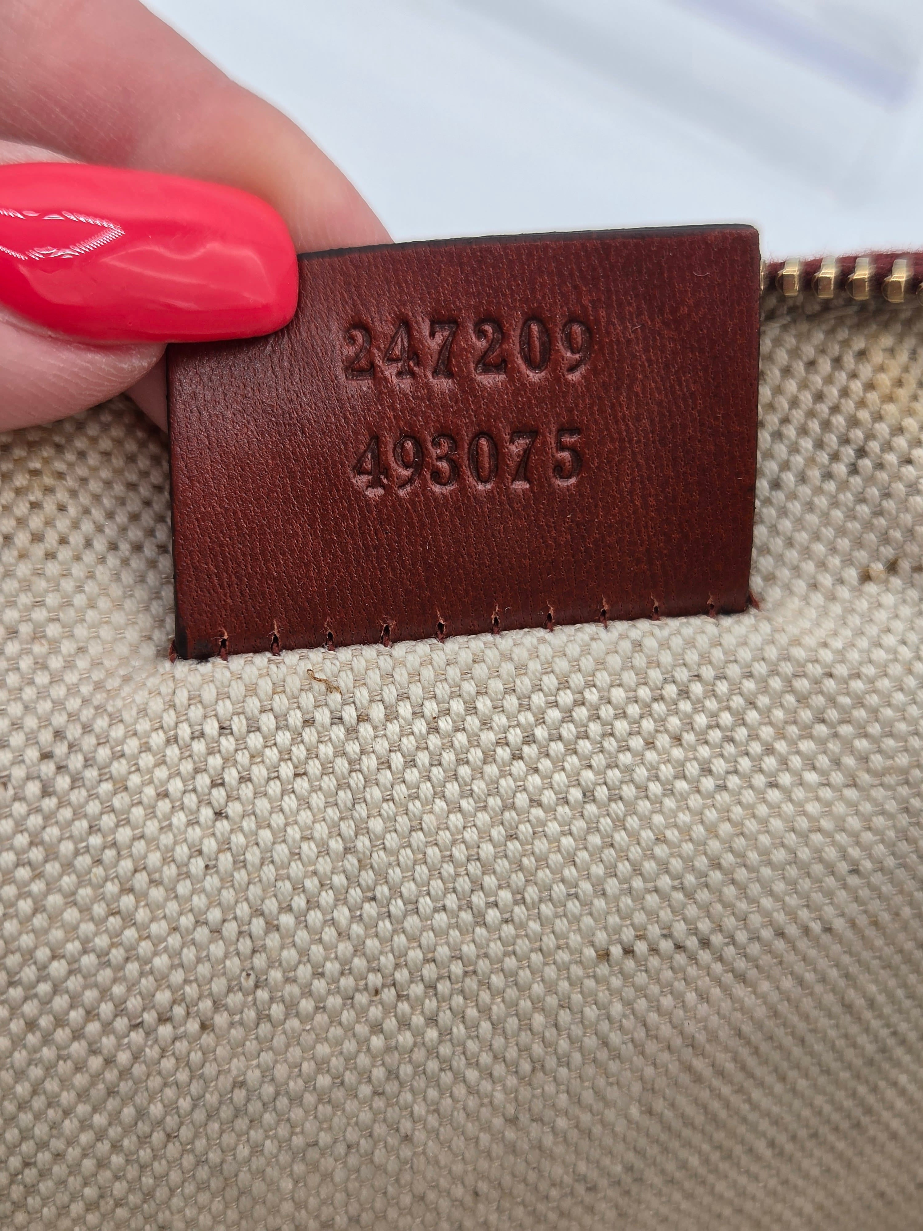 Close-up of a beige woven fabric item with a brown leather tag stamped with the numbers 247209 and 498075, held by a hand with bright pink nail polish.
