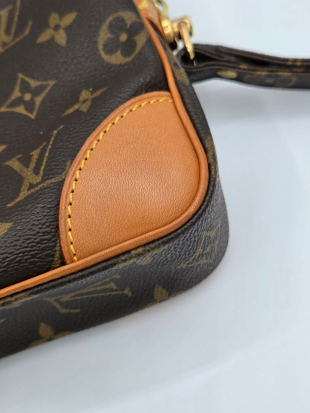 Close-up of a Louis Vuitton monogram handbag corner showing dark brown coated canvas with tan leather trim and gold-tone hardware on a white background.