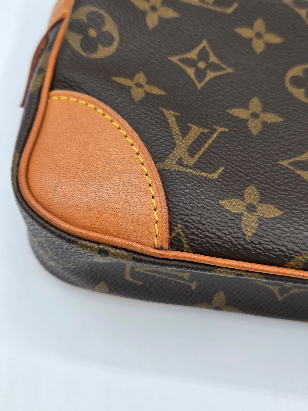 Close-up of a Louis Vuitton monogram canvas pouch or bag corner with tan leather trim and yellow contrast stitching on a white background.