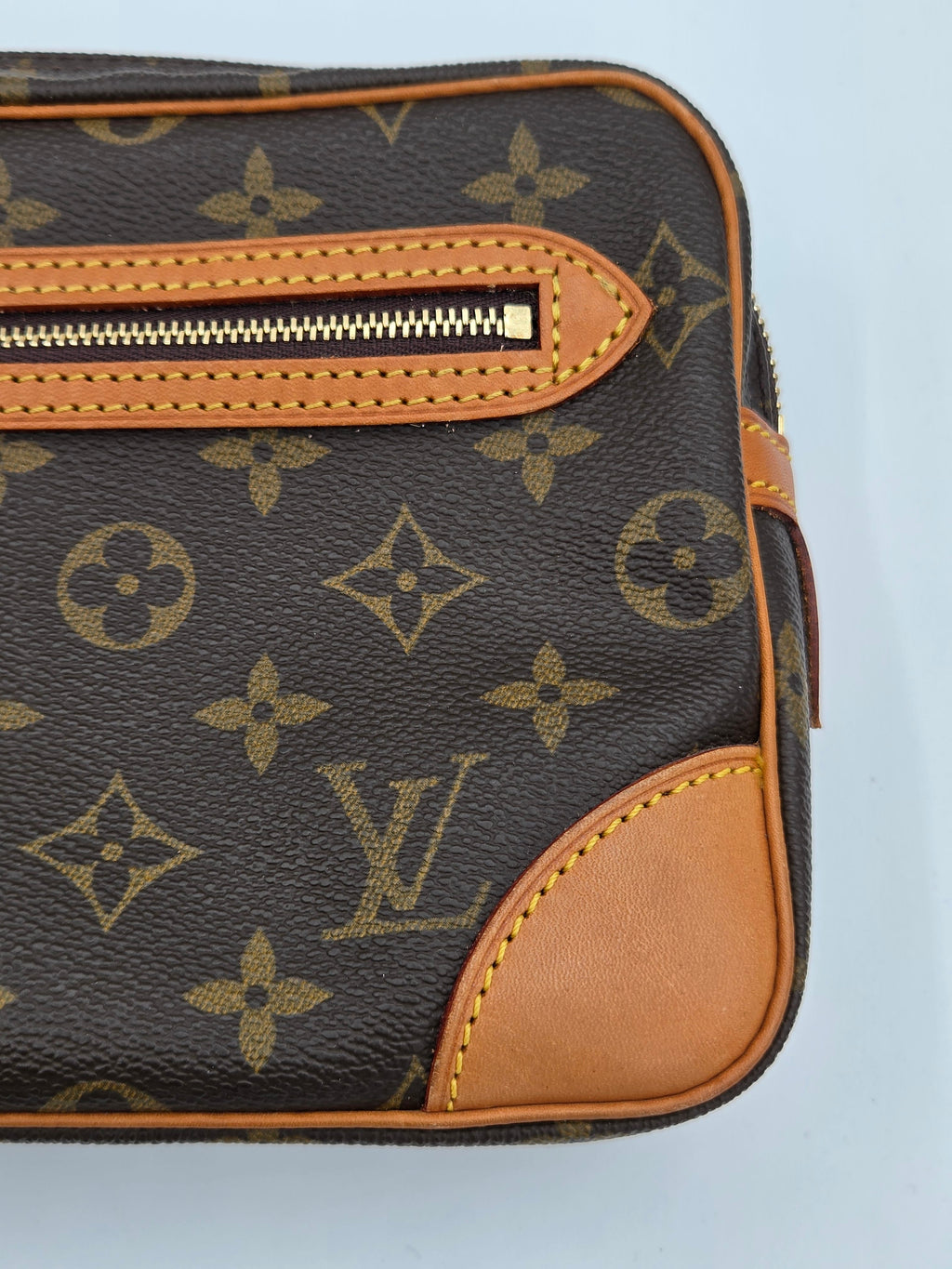 Close-up of a Louis Vuitton monogram canvas pouch or bag corner with tan leather trim, a gold zipper, and the Louis Vuitton LV monogram pattern on a light blue background.