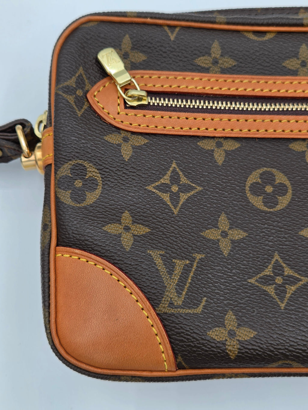 Close-up of a Louis Vuitton monogram canvas pouch or small bag, showing the dark brown coated canvas with tan monogram motifs, tan leather trim, a gold zipper pull, and a tan leather corner protector on a light gray background.