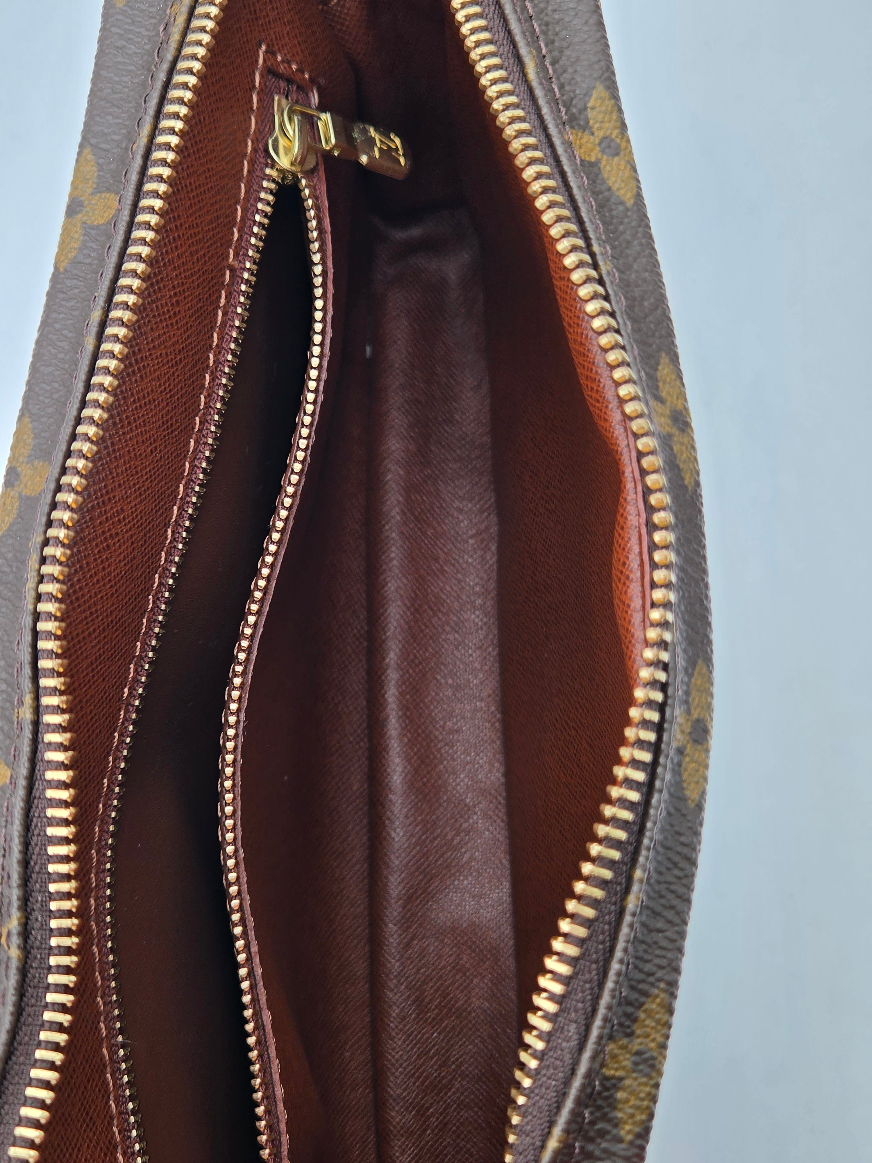 Close-up of the interior of a brown monogram handbag, showing a dark brown leather lining, gold-tone zipper pulls, and the signature patterned exterior material against a light blue background.