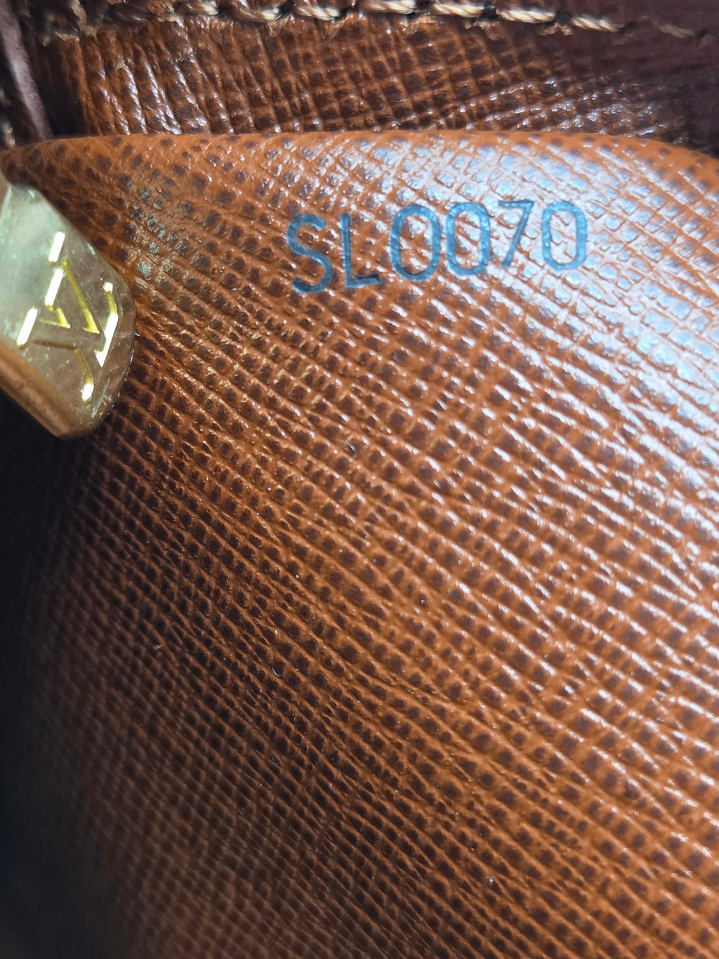 Close-up of a brown textured leather interior with embossed blue lettering reading 'SL0070' and a small gold-toned hardware piece visible on the left.