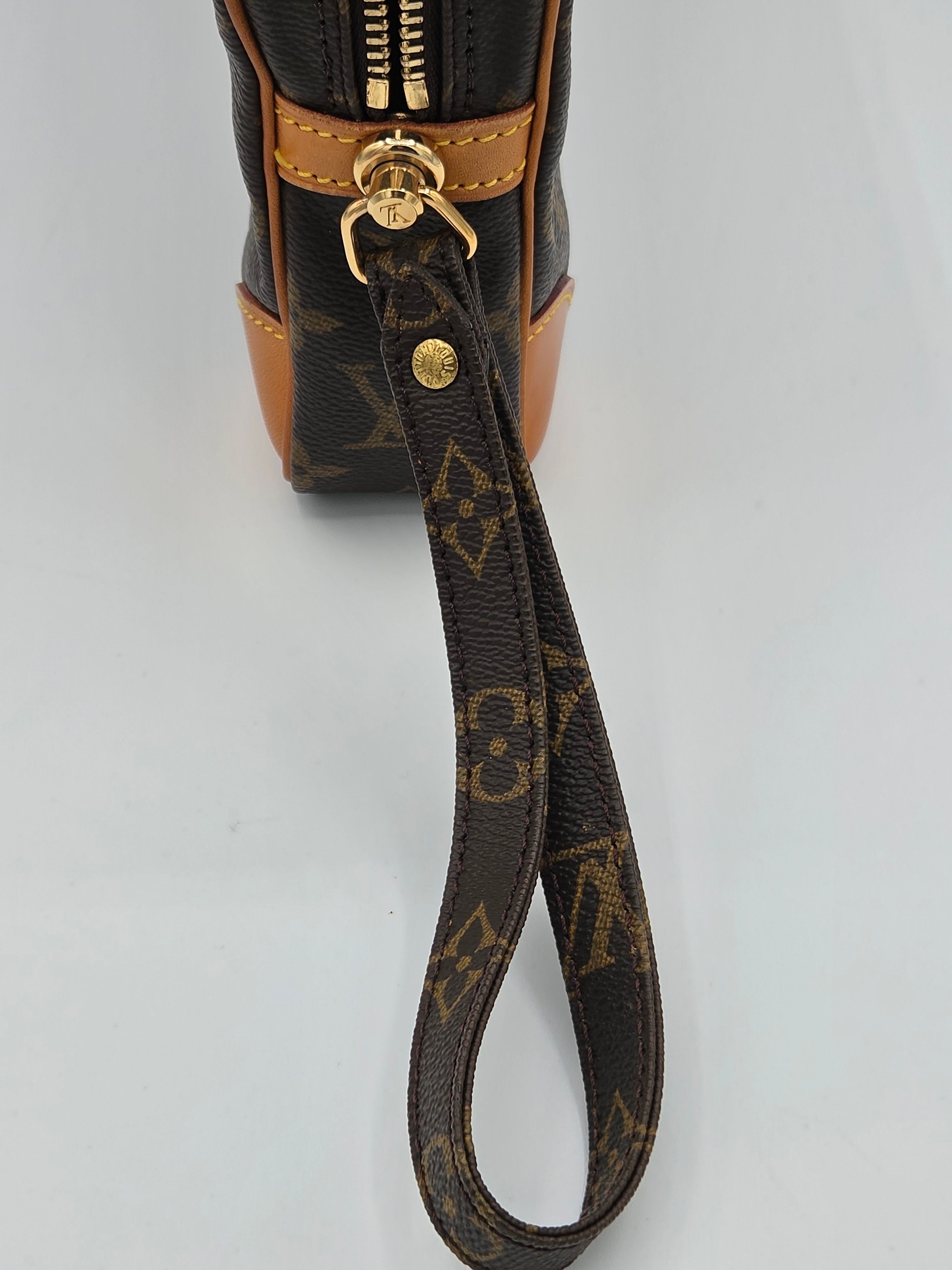 Close-up of a Louis Vuitton monogram wristlet bag showing the brown canvas body, tan leather trim, gold-tone hardware, and the attached monogram wrist strap on a light gray background.