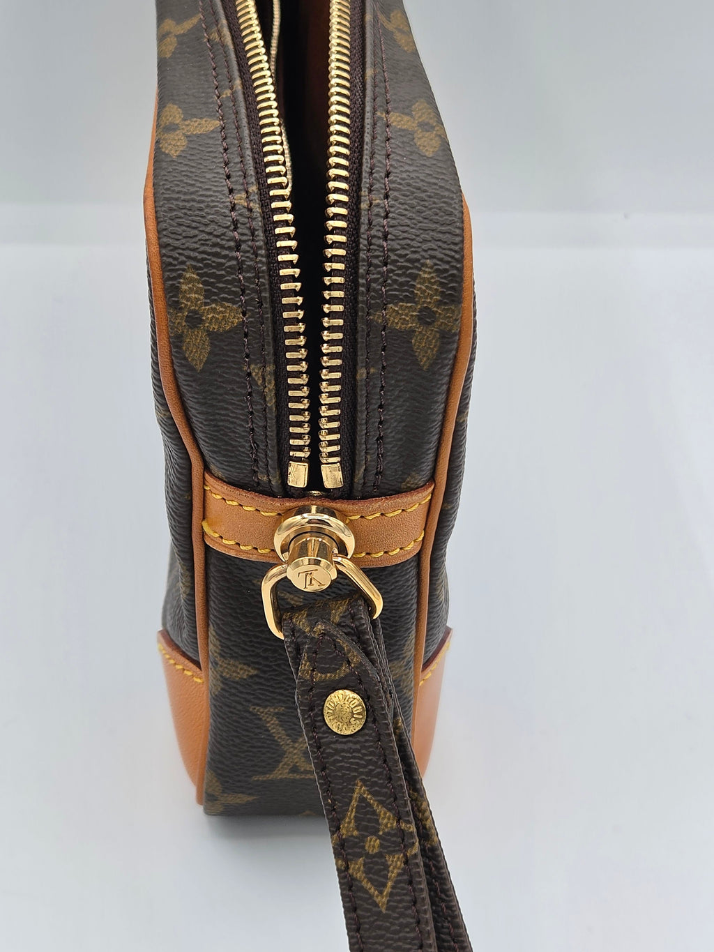 Close-up of a Louis Vuitton monogram canvas bag with tan leather trim and gold-tone hardware, showing the zipper and wristlet strap detail.