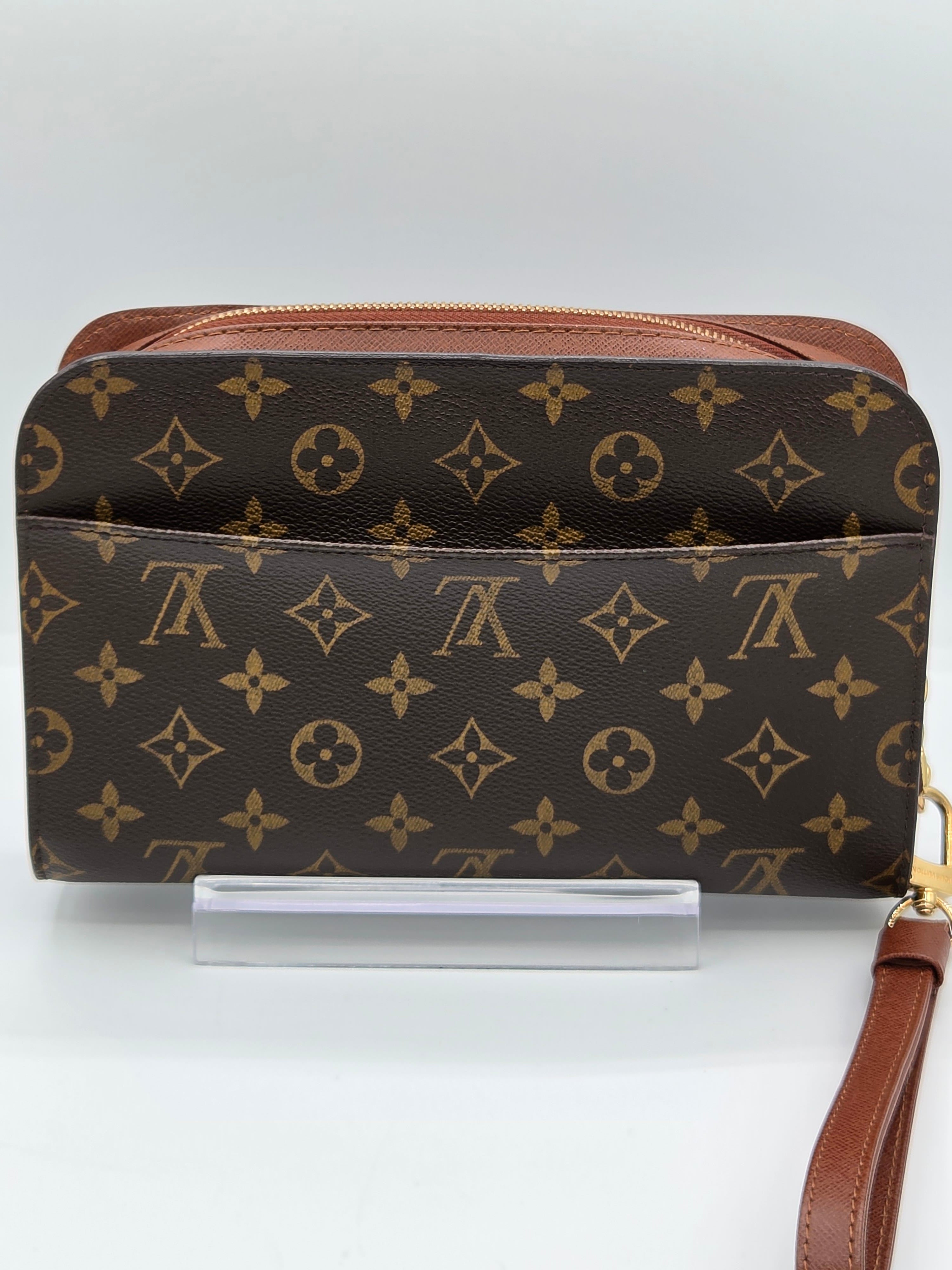 A Louis Vuitton monogram canvas zip-around wallet or clutch with a brown leather wrist strap, displayed on a clear acrylic stand against a light gray background.