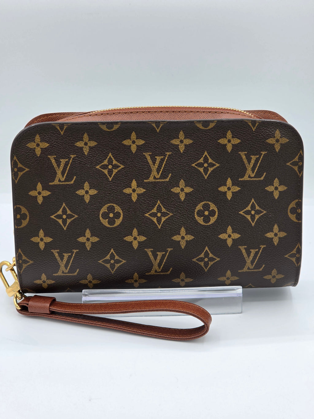 A Louis Vuitton monogram canvas wristlet pouch with a brown leather wrist strap and gold-tone hardware, displayed on a clear stand against a light gray background.