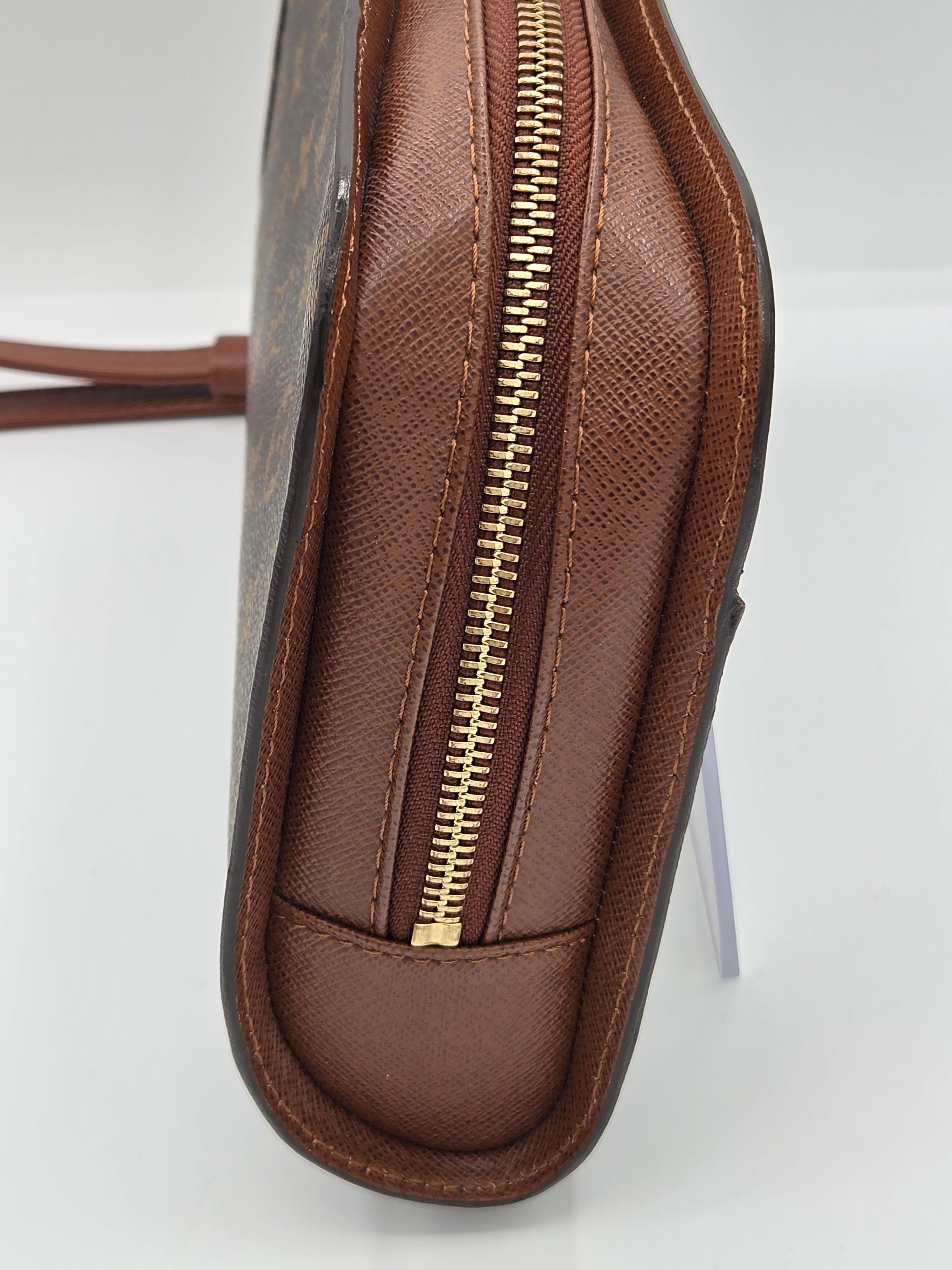 Close-up of a brown textured leather bag corner showing a gold-tone zipper and a small portion of the monogram canvas and strap.